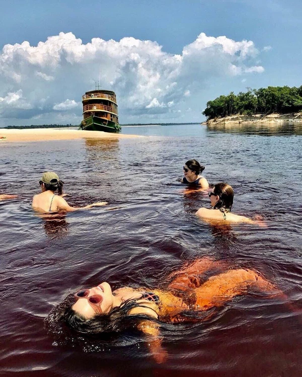 Dunkel wie Tee, warm wie eine Badewanne, niemand bleibt lange am Ufer. Vier Reisende schwimmen im dunkel tanninhaltigen Wasser des Rio Negro auf einer Amazonas-Kreuzfahrt in Brasilien, im Hintergrund ein klassisches mehrstöckiges Holzflussschiff vor einer weißen Sandbank
