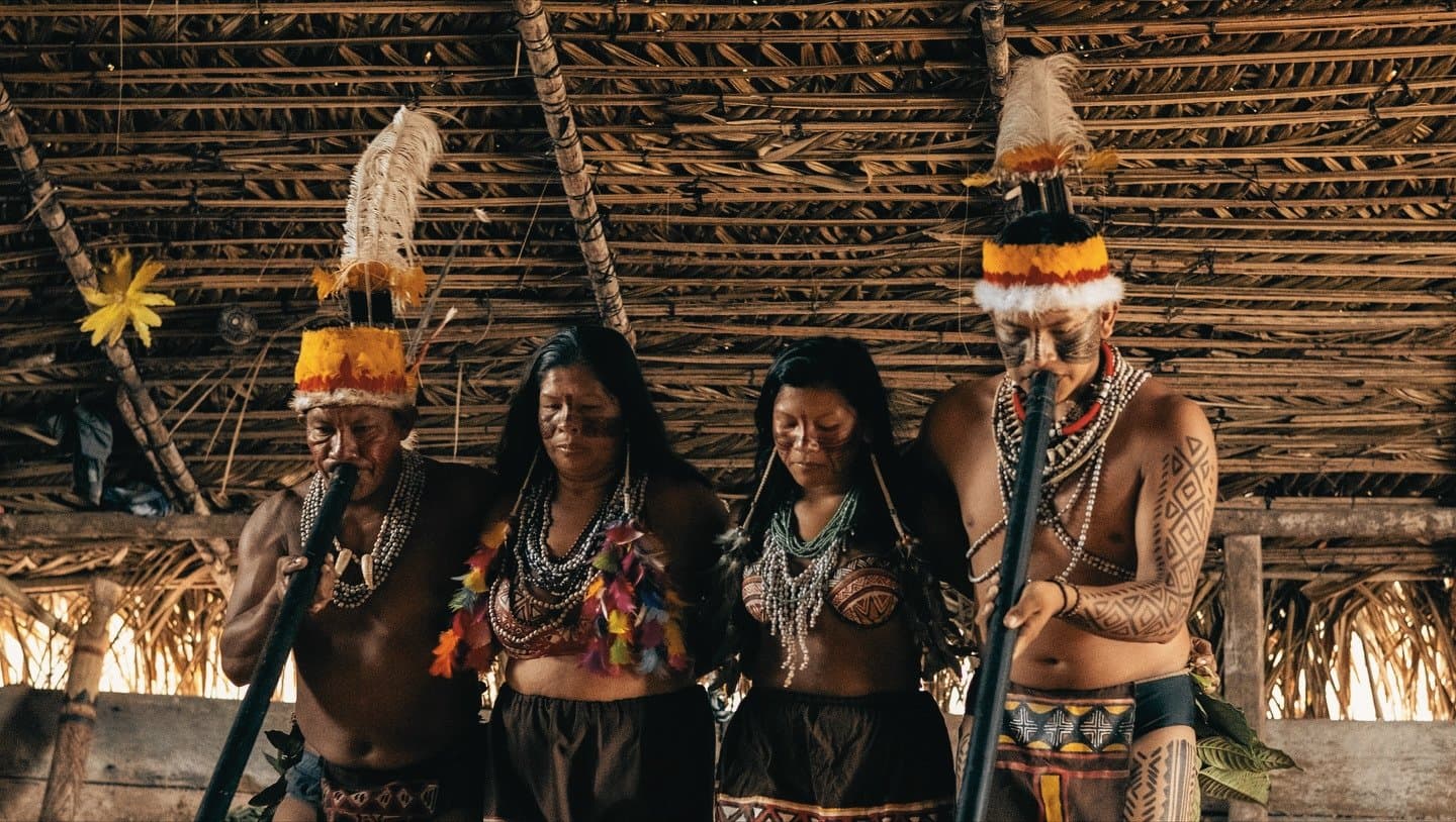 Gesänge, die hier schon immer erklungen sind, und es immer sein werden. Vier Mitglieder einer indigenen Gemeinschaft in traditionellen Federkopfschmuck, Körperbemalung und Perlenketten führen eine Zeremonie in einer Maloca mit Strohdach durch