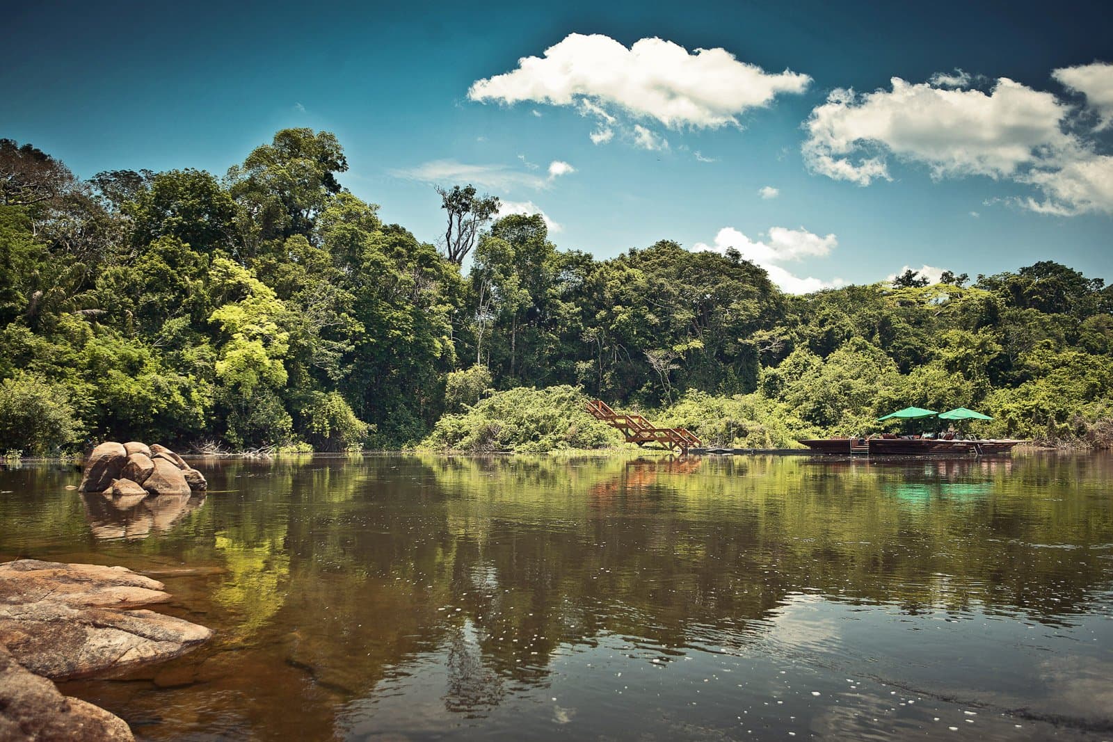 La rivière, dernier couloir avant la forêt. Le reste peut attendre. Photo: Samuel Melim (c) Une rivière aux eaux sombres et calmes reflète la canopée dense de la forêt amazonienne ; des rochers granitiques longent la rive proche tandis qu'un escalier en bois et deux parasols verts ombragent un ponton sur la rive opposée — point d'arrivée pour l'observation des oiseaux en forêt amazonienne