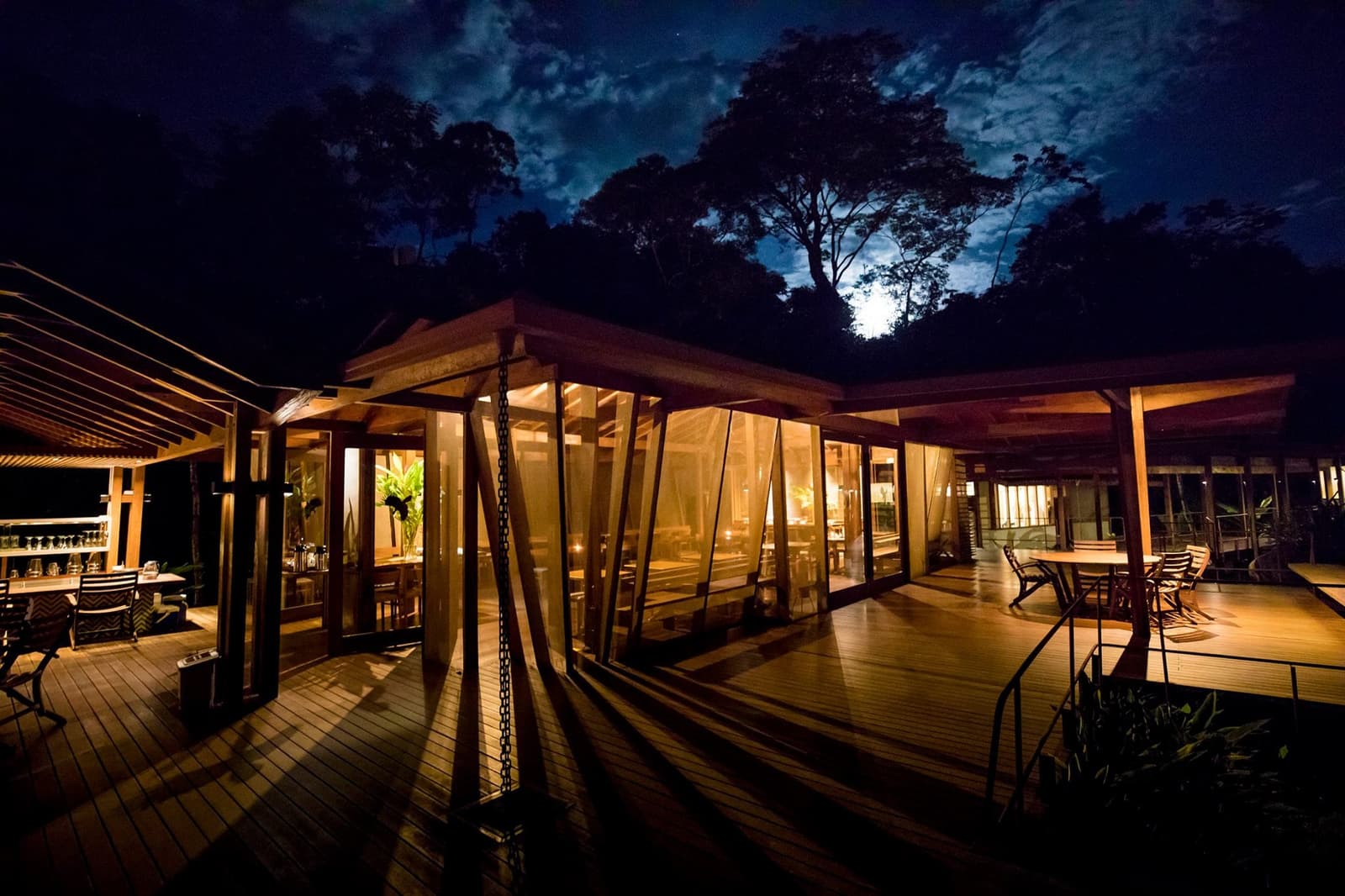 Le dîner est fini, la forêt dort, et le bar est encore ouvert. On a connu pire. Le restaurant et la terrasse en bois du Cristalino Jungle Lodge illuminés la nuit, lumière ambrée filtrant à travers les parois vitrées, silhouettes des arbres sous un ciel de lune