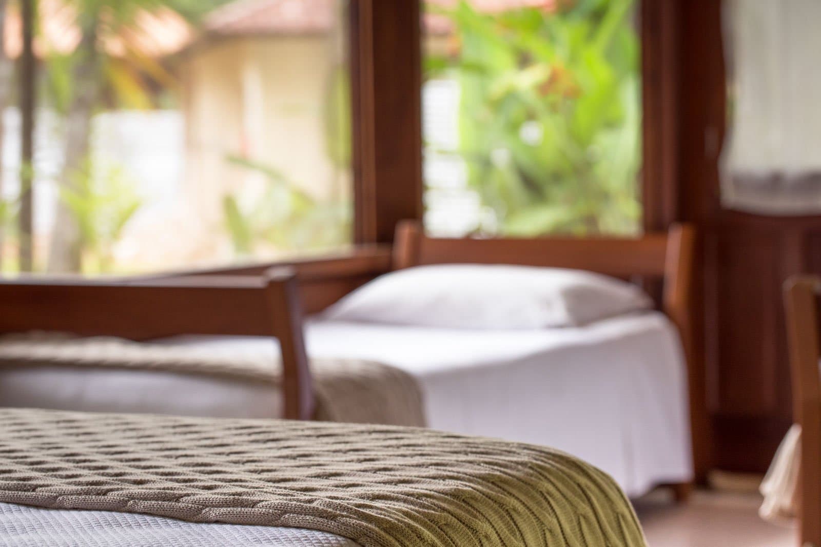 Draps bien frais, un plaid douillet, et la forêt à portée de regard. Photo: | SamuelMelim Photography Deux lits à cadre en bois avec un plaid vert tricoté dans une chambre de lodge, grandes fenêtres donnant sur la végétation tropicale — hébergement confortable pour l'observation des oiseaux en Amazonie