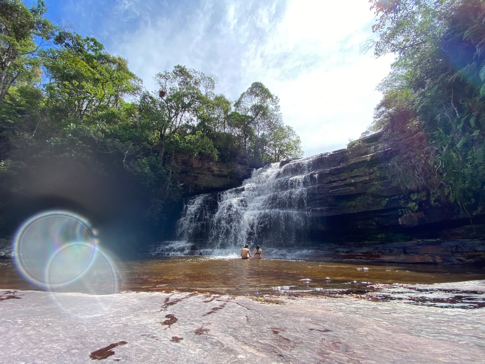 Kühles Wasser nach einem langen Morgen auf dem Trail, besser wird's nicht. Zwei Schwimmer waten in einem natürlichen Pool am Fuß eines breiten, mehrschichtigen Wasserfalls, der über Sandsteinplatten fällt, umgeben von grünen Bäumen im Chapada Diamantina