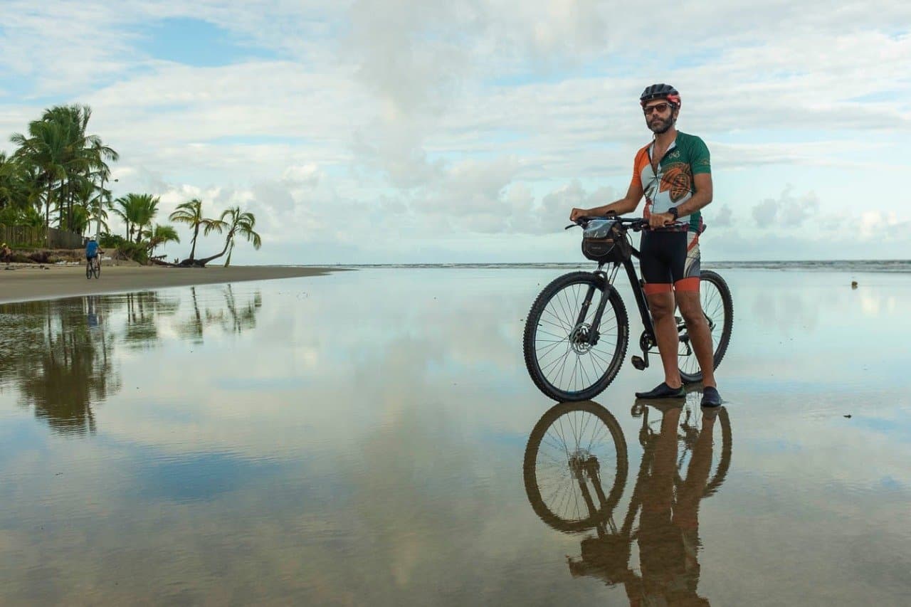 Nasser Sand, ein perfekter Spiegel, und noch viele Kilometer vor sich. Ein Radfahrer in grün-orangenem Trikot hält sein Mountainbike auf einem spiegelglatten, nassen Strand; Palmen und bewölkter Himmel spiegeln sich im flachen Wasser, im Hintergrund ist ein weiterer Radfahrer zu sehen.