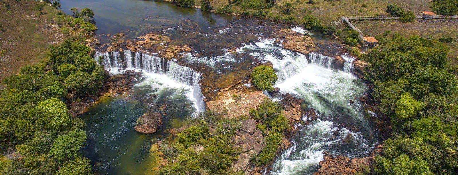 Zwölf Kilometer zu Fuß, und dann biegt man um die Ecke und sieht das hier. Luftaufnahme eines breiten, hufeisenförmigen Wasserfalls auf einem felsigen Plateau, umgeben von Cerrado-Vegetation, mit einem kleinen Aussichtspunkt am rechten Ufer auf einer Jalapão-Trekkingroute