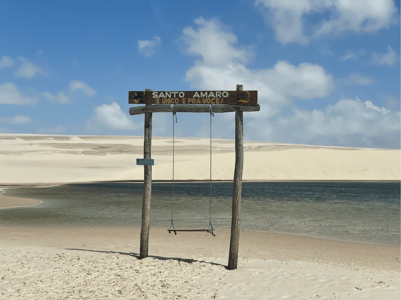 Santo Amaro hat ein Schild aufgestellt, nur für den Fall, dass man es vergisst. Ein rustikales Holzschaukelgestell mit dem Schild 'Santo Amaro é único e pra você' steht auf weißem Sand neben einer ruhigen Lagune mit weitläufigen weißen Dünen im Hintergrund