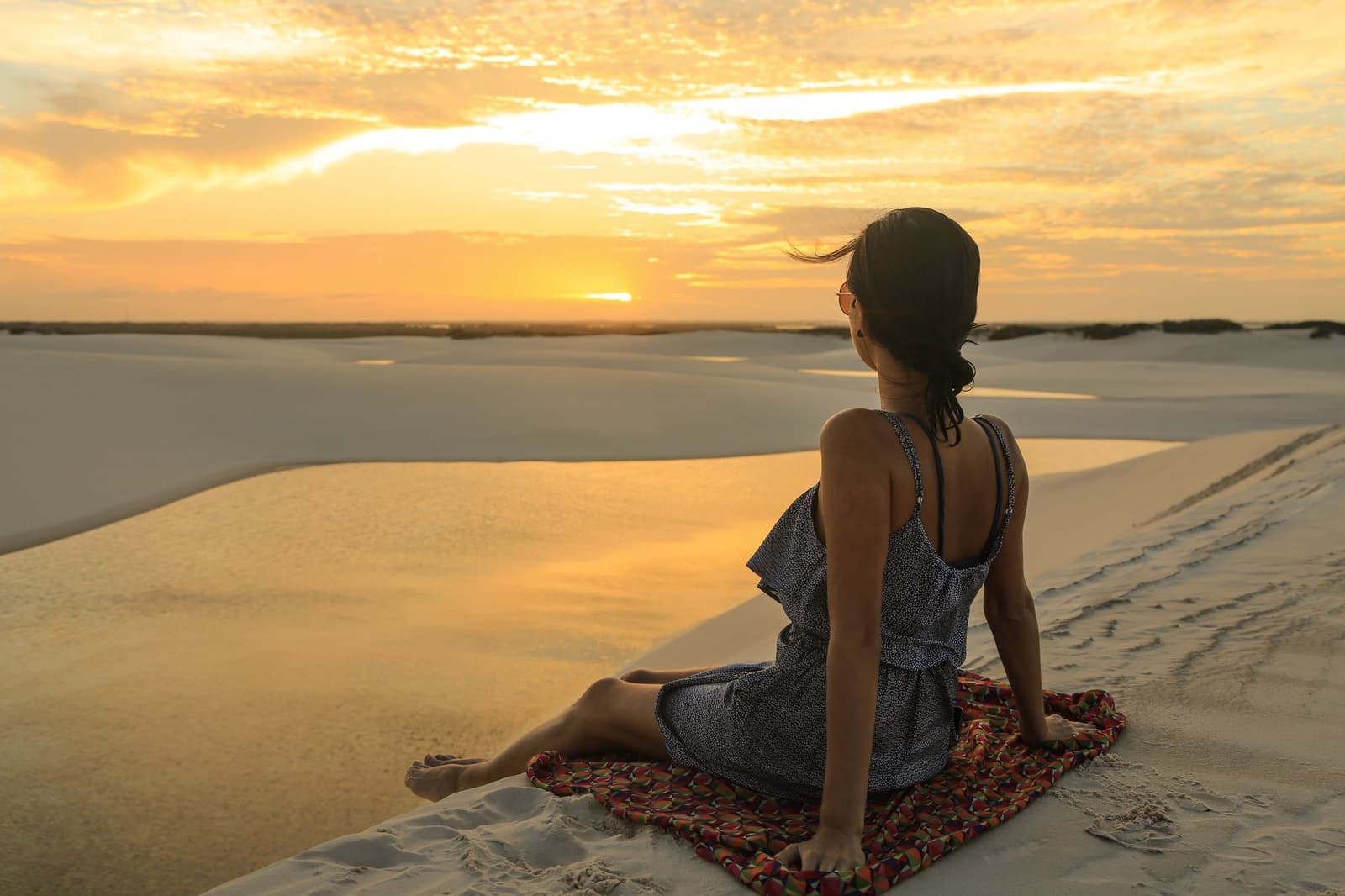 Für manche Aussichten legt man sich einfach hin und bleibt. Eine Frau sitzt auf einer bunten Decke auf einer weißen Sanddüne und blickt auf einen leuchtend goldenen Sonnenuntergang über der Dünenlandschaft der Lençóis Maranhenses