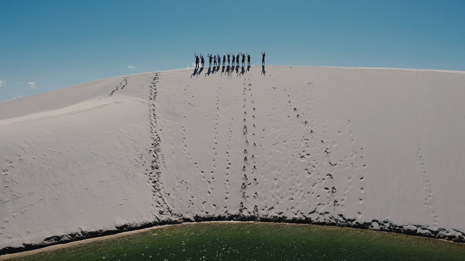 Oben angekommen, die Düne trägt noch jeden Schritt als Erinnerung. Luftaufnahme einer Gruppe von Wanderern, die mit erhobenen Armen auf dem Kamm einer riesigen weißen Sanddüne beim Lençóis-Maranhenses-Trekking stehen, mit einer grünen Lagune am Fuß und blauem Himmel darüber
