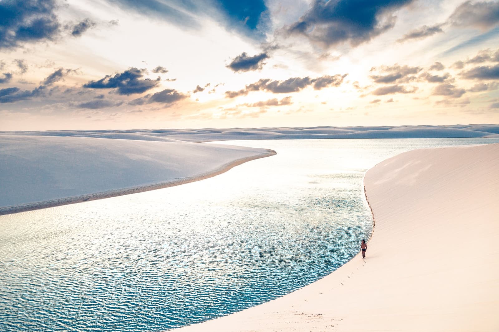 Weiß, blau, weit. Mehr braucht dieser Ort nicht. Eine einsame Wanderin läuft entlang einer spiegelnden Süßwasserlagune, eingebettet zwischen weißen Sanddünen unter einem goldenen Abendhimmel im Lençóis Maranhenses