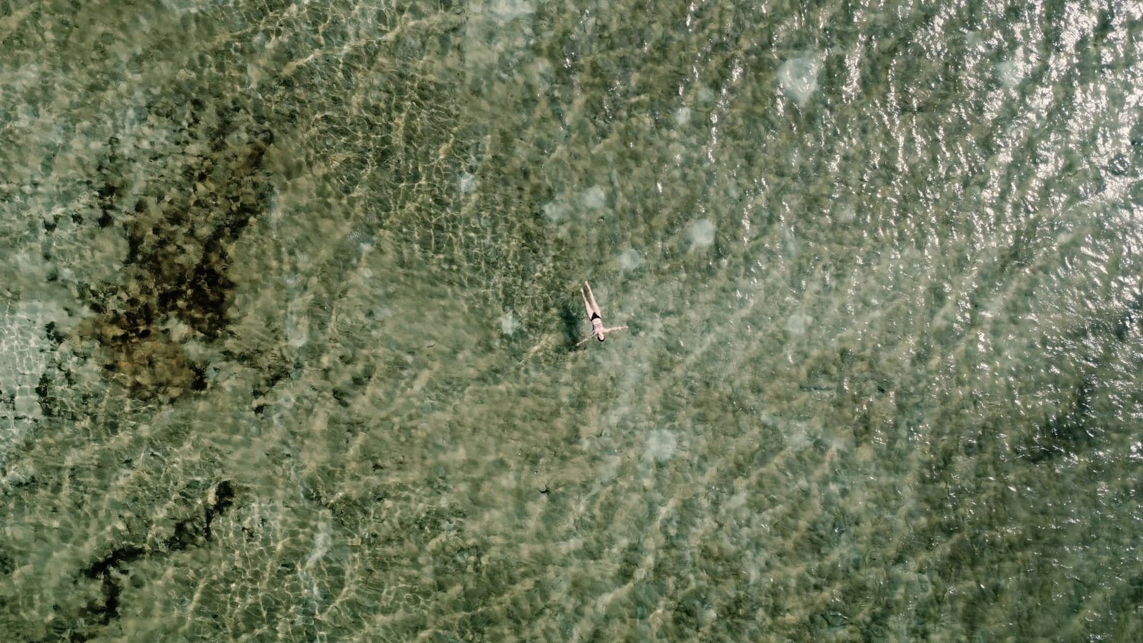 Allein im Wasser, und das fühlt sich genau richtig an. Luftaufnahme einer einzelnen Person, die in flachem, kristallklarem grünen Wasser über einem felsigen Untergrund auf dem Rücken schwimmt