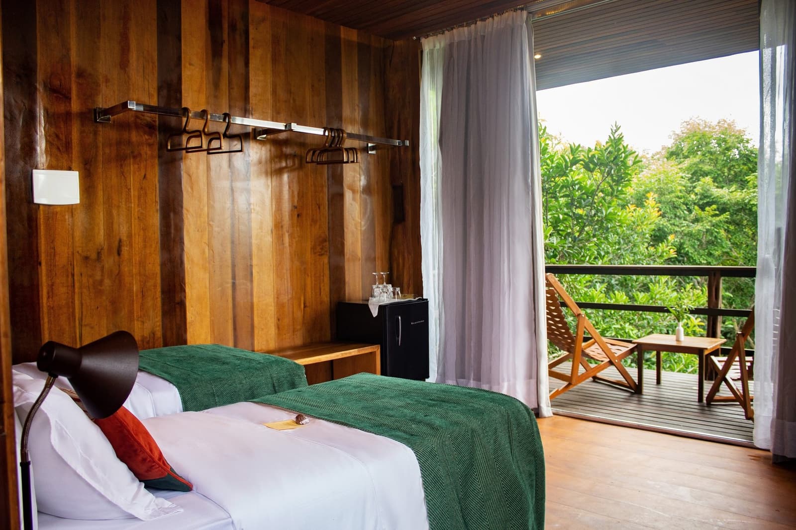 Vorhang auf, Regenwald. So sieht der Ausblick vom Bett aus. Holzvertäfeltes Gästezimmer im Mirante do Gavião mit ordentlich gemachtem Bett, Minikühlschrank und offenen Glastüren zu einem privaten Balkon mit Blick in den dichten Amazonas-Regenwald