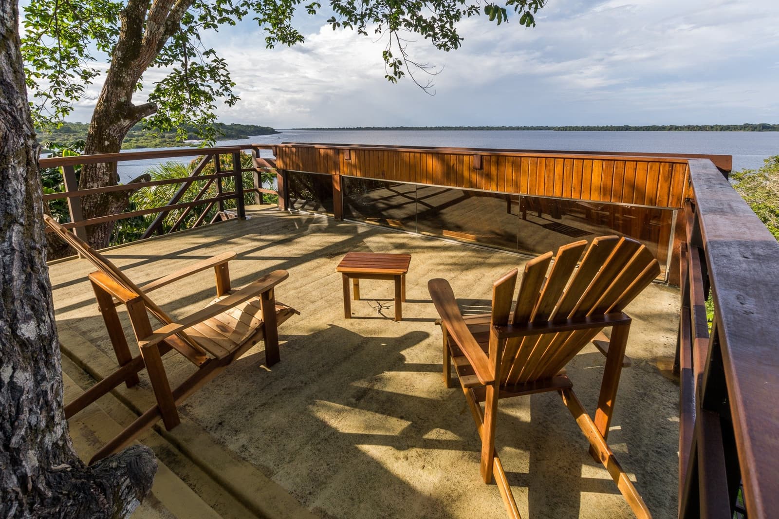 Einfach sitzen, zuschauen, wie der Fluss die Zeit angibt. Zwei Holzliegestühle und ein kleiner Tisch auf einer offenen Holzterrasse des Mirante do Gavião, mit Blick auf einen breiten Schwarzwasserfluss und dichten Regenwald