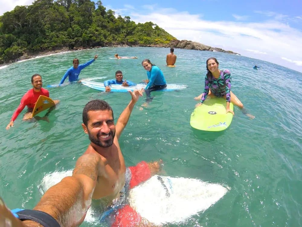 Cinco pranchas, sorrisos à vontade, e ninguém com pressa de sair do mar. Um instrutor sorridente tira uma selfie no mar com um grupo de alunos durante aulas de surf em Itacaré, todos segurando pranchas coloridas nas águas verde-turquesa com um promontório coberto de mata ao fundo.