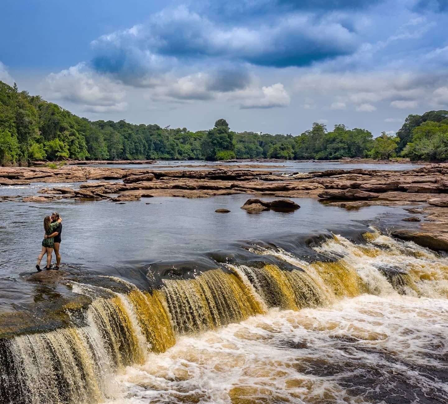 Manche Momente plant man nicht, der Fluss übernimmt das einfach. Ein Paar umarmt sich auf einem Felsen neben einem breiten Wasserfall inmitten tropischen Urwalds auf einer Amazon Jungle Cruise durch Brasilien, unter dramatisch bewölktem Himmel