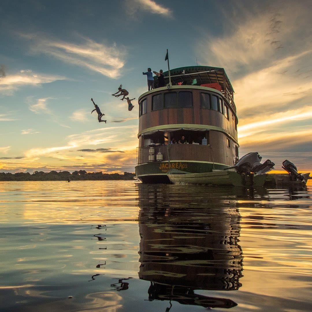 Am Ende eines guten Tages ist der Sprung ins Wasser einfach die richtige Entscheidung. Gäste springen bei Sonnenuntergang vom Oberdeck des Flussboots Jacareagil in den spiegelglatten Fluss auf einer Amazonas-Flusskreuzfahrt in Brasilien, goldenes Licht spiegelt sich im schwarzen Wasser