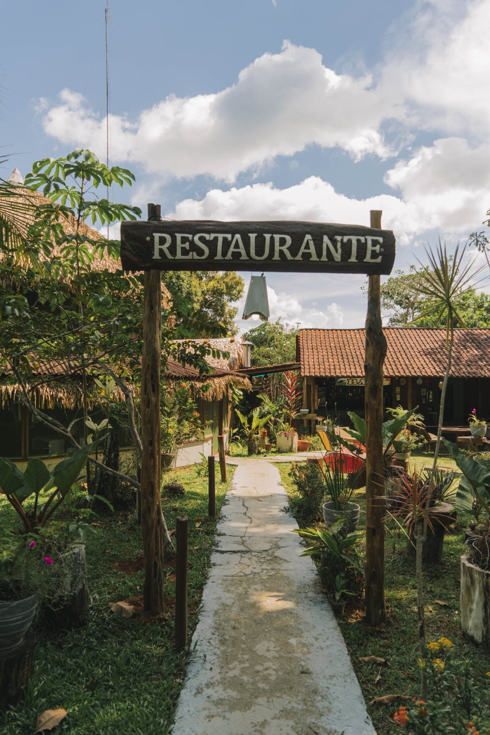Der Weg zum Essen ist hier schon halb das Erlebnis. Ein rustikales Holzschild mit der Aufschrift 'Restaurante' überspannt einen Steinweg, der durch einen üppigen tropischen Garten zum Freiluft-Restaurant des Amazon Turtle Lodge führt