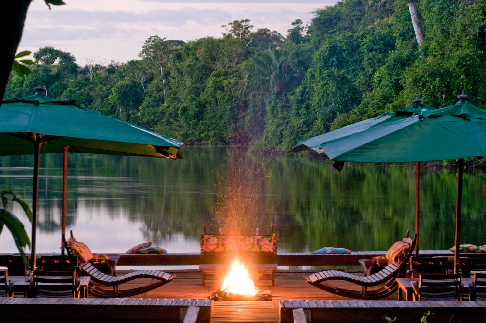 Feu de camp, eau dormante, forêt toute proche. Exactement ce qu'il faut après une journée de sentiers. Photo: Luis Gomes Un feu de camp allumé sur une terrasse en bois au crépuscule, entouré de chaises longues rembourrées et de parasols verts, face à une rivière calme reflétant la forêt tropicale dense — un cadre idéal pour l'observation des oiseaux en forêt amazonienne.