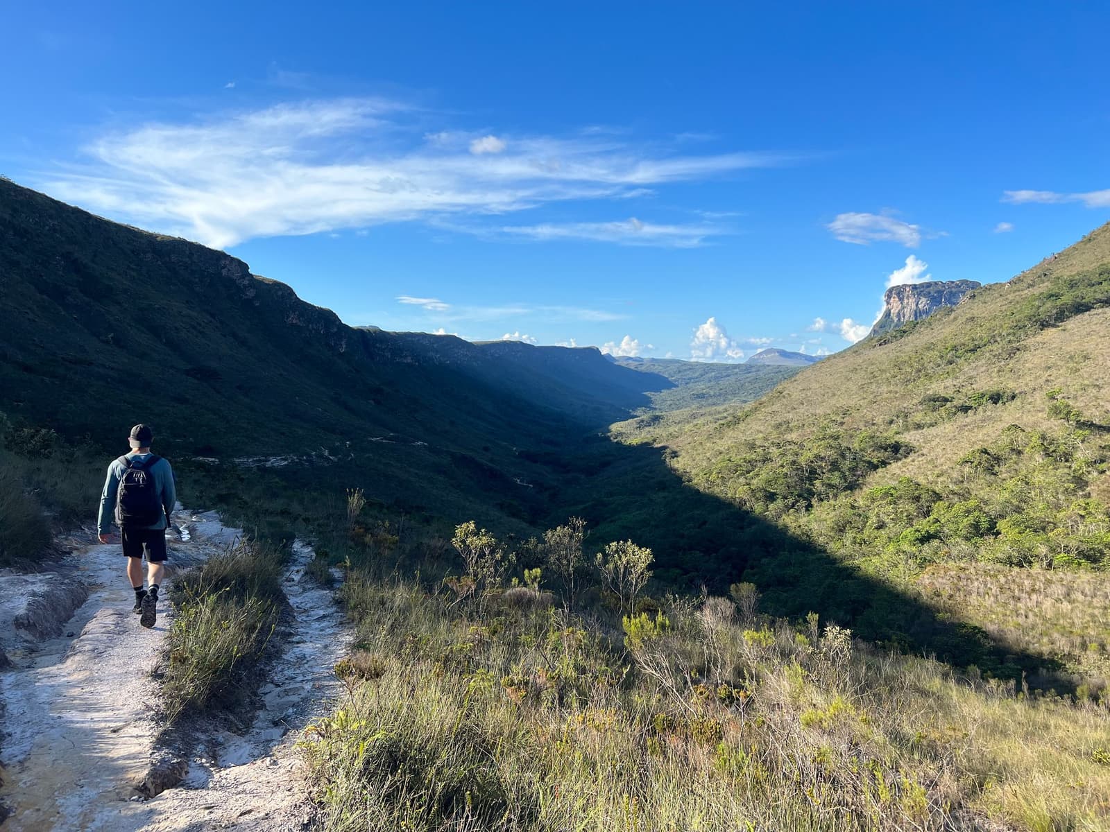 Der Weg geht weiter, und irgendwie auch du. Ein Wanderer mit Rucksack geht auf dem Chapada Diamantina Trek einen felsigen Pfad entlang, während sich ein weites grünes Tal zwischen Sandsteinfelsen unter blauem Himmel erstreckt