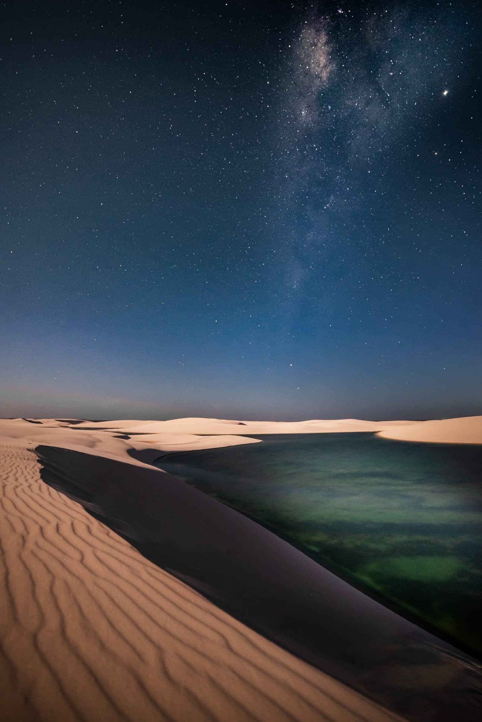 Sanddünen, eine stille Lagune und die ganze Galaxie für sich allein. Die Milchstraße wölbt sich über weiße Sanddünen und eine ruhige türkisfarbene Lagune bei Nacht im Lençóis Maranhenses Full Itinerary Sterngucken-Erlebnis