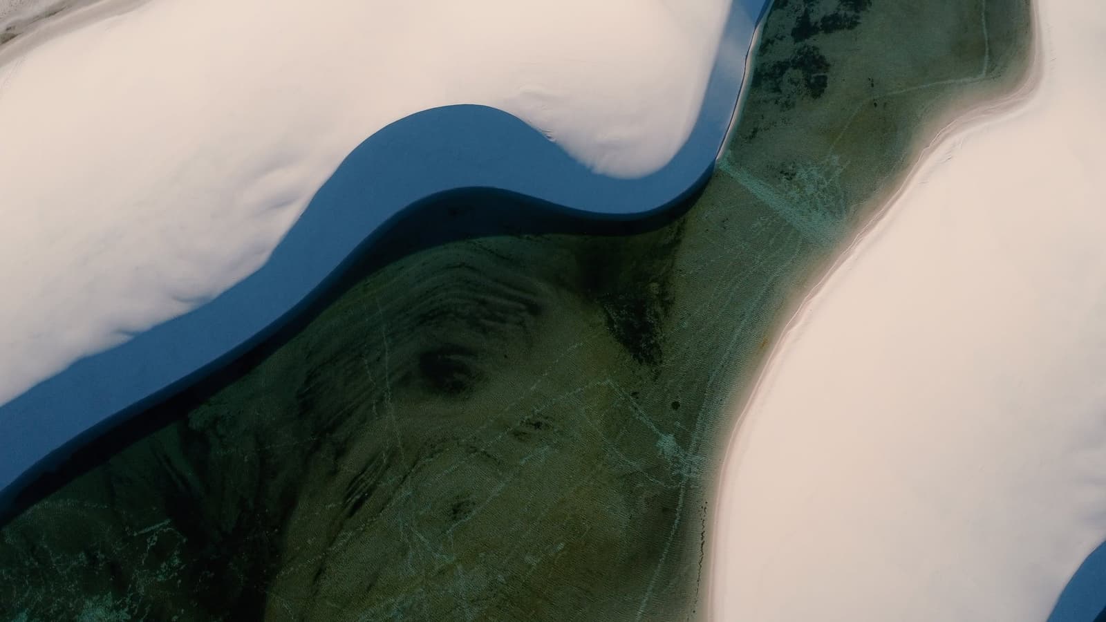 Aus der Vogelperspektive wirkt es, als würden die Dünen die Landschaft Stück für Stück verschlucken. Luftaufnahme einer tiefblauen Lagune, die sich durch weiße Sanddünen zieht, mit grüner Unterwasservegetation im klaren Wasser