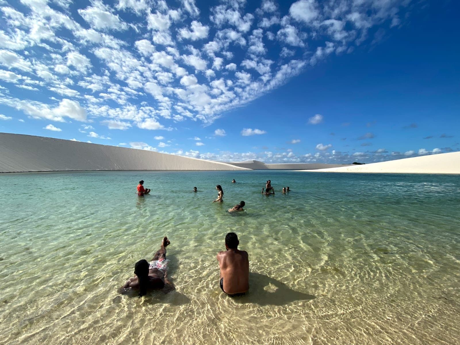 Regenwasser, weiße Dünen und kein Handtuch in Sicht. Genau so soll das sein. Mehrere Personen schwimmen und waten in einer glasklaren türkisfarbenen Süßwasserlagune bei einer Wanderung durch die Lençóis Maranhenses, mit sanften weißen Sanddünen am gegenüberliegenden Ufer unter einem blauen Himmel mit kleinen Wolken