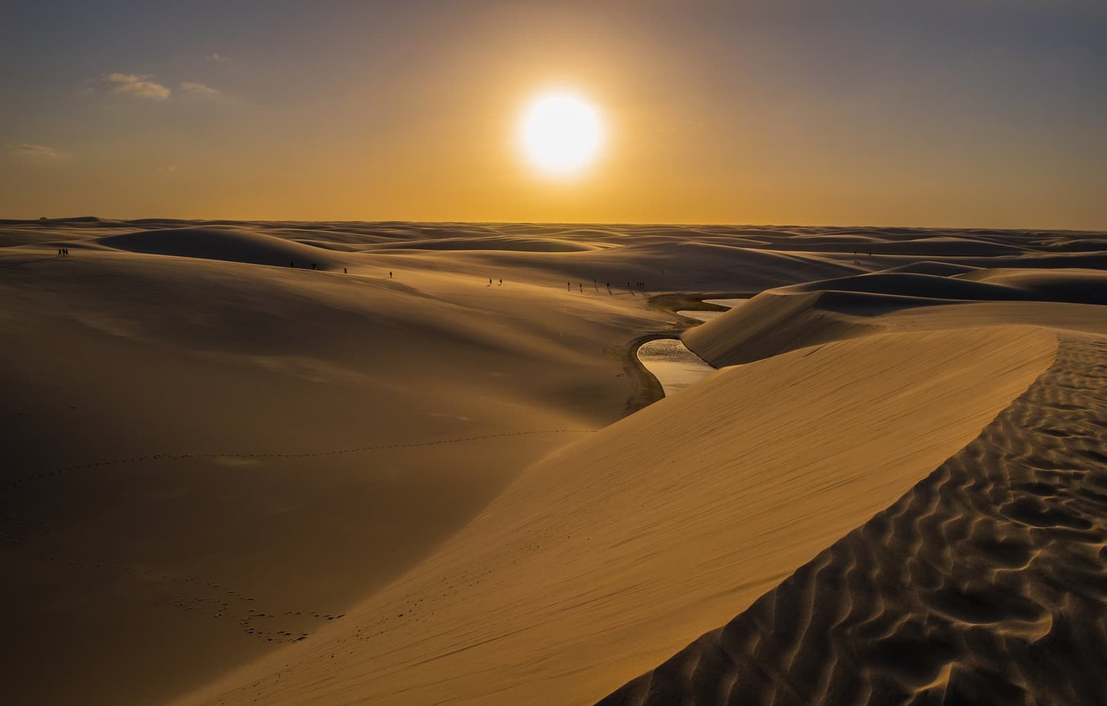 Eine Wüste, die sich mit Wasser füllt. Die Lençóis Maranhenses spielen nach eigenen Regeln. Riesige goldene Sanddünen der Lençóis Maranhenses erstrecken sich bei Sonnenuntergang bis zum Horizont, mit einer kleinen Süßwasserlagune zwischen den Dünen und winzigen Silhouetten von Wanderern in der Ferne