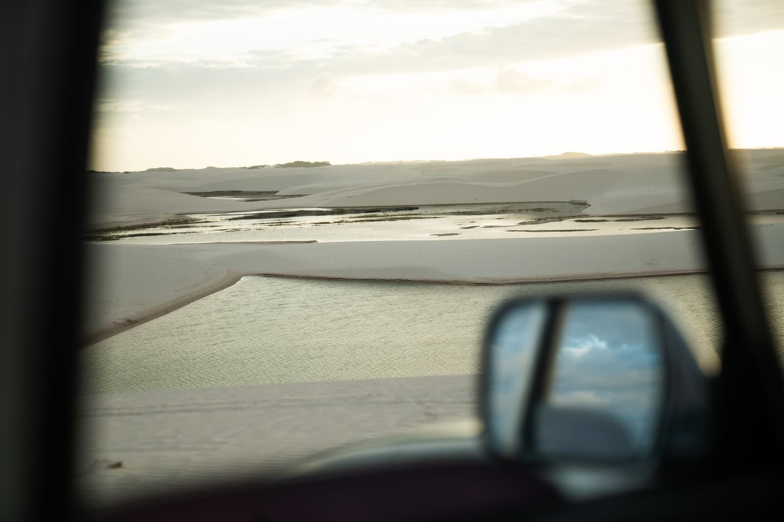 Noch im Wagen, und die Dünen füllen schon das ganze Fenster. Blick durch das Fenster eines Geländewagens auf weiße Sanddünen und flache grüne Lagunen, die sich beim Lençóis-Maranhenses-Trek von Atins bis zum Horizont erstrecken, mit dem Seitenspiegel, der einen bewölkten Himmel widerspiegelt
