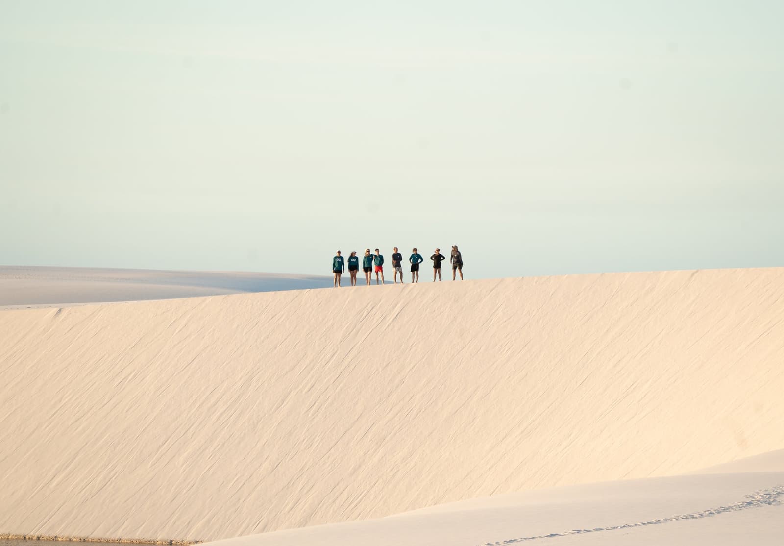 Perchés sur la dune, ils mesurent enfin l'échelle de l'endroit. Sept randonneurs debout sur la crête d'une immense dune de sable blanc lors d'un trek dans les Lençóis Maranhenses, avec un ciel pâle et un paysage de dunes s'étendant à perte de vue derrière eux