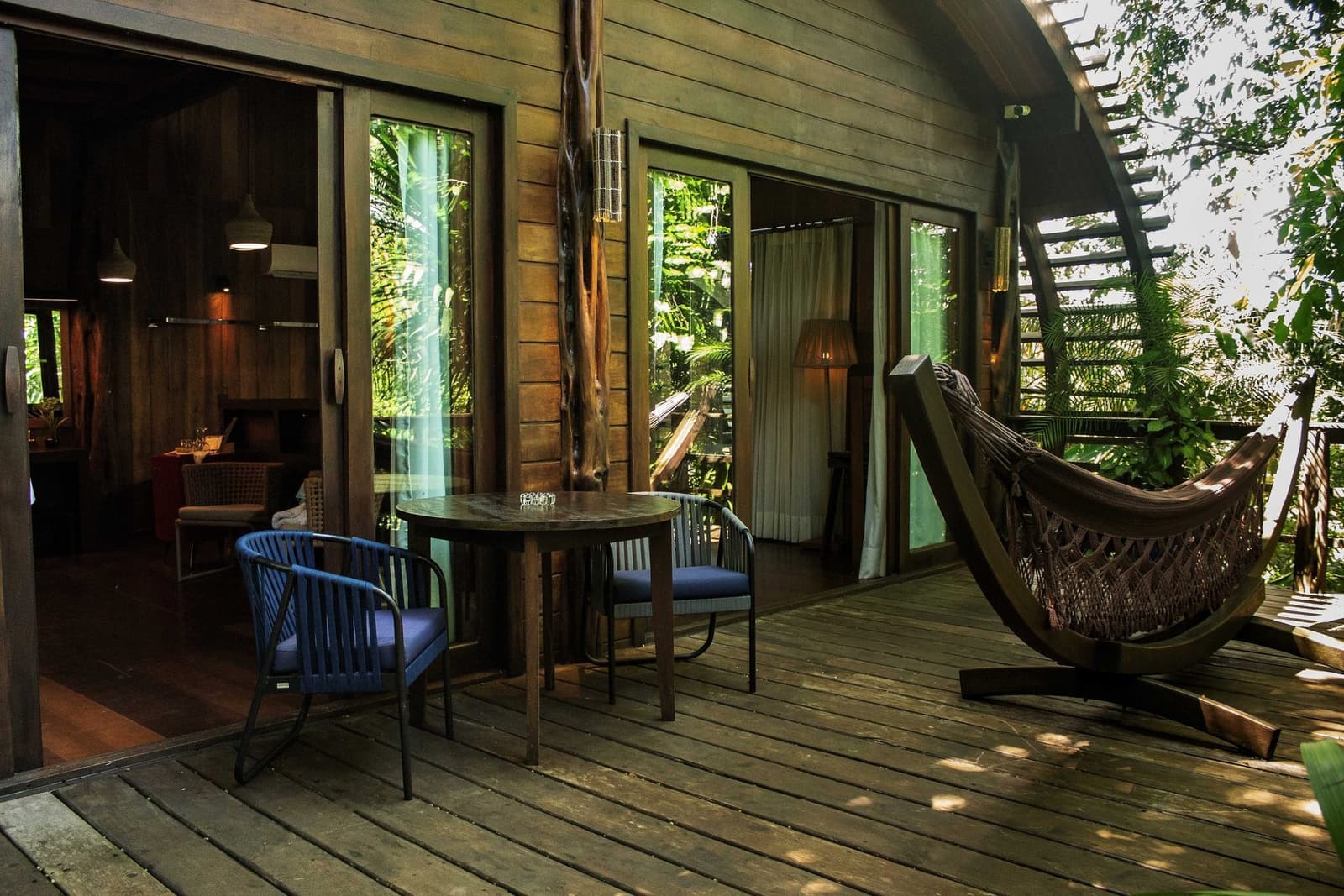 Wo der Regenwald so nah ist, braucht man kein Fenster aufzumachen. Eine Holzterrasse im Mirante do Gavião mit einem runden Tisch, zwei gepolsterten Stühlen, einer geflochtenen Hängematte auf einem geschwungenen Ständer und Glasschiebetüren, die in ein stimmungsvoll beleuchtetes Zimmer inmitten üppigen Regenwaldes führen