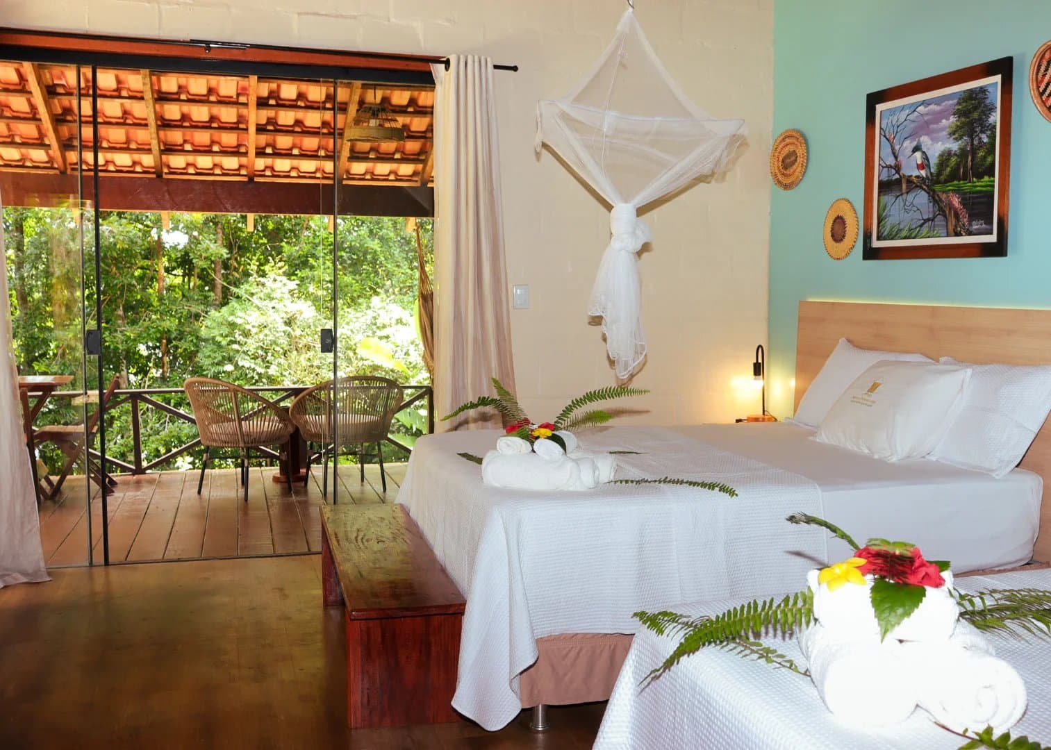 Frische Bettwäsche, ein Moskitonetz, und der Regenwald direkt hinter der Glastür. Innenraum eines Zimmers in einer Amazon-Boutique-Lodge mit zwei weiß bezogenen Betten, gerollten Handtüchern und tropischen Blüten als Deko, einem hängenden Moskitonetz, warmem Nachtlicht, lokaler Kunst an der Wand und Glasschiebetüren, die auf einen Holzbalkon mit Rattanstühlen und dichtem Regenwald führen