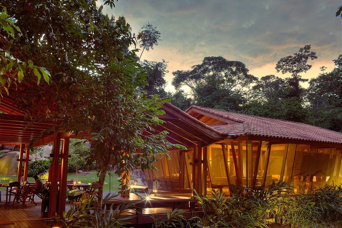Bougies allumées, forêt toute proche, rivière juste derrière. Le lodge sait comment finir une journée. Photo: Samuel Melim Le Cristalino Jungle Lodge illuminé de l'intérieur au crépuscule, son pavillon de bois et de verre entouré de la canopée dense de la forêt amazonienne et d'un jardin tropical, lanternes allumées sur la terrasse en bois
