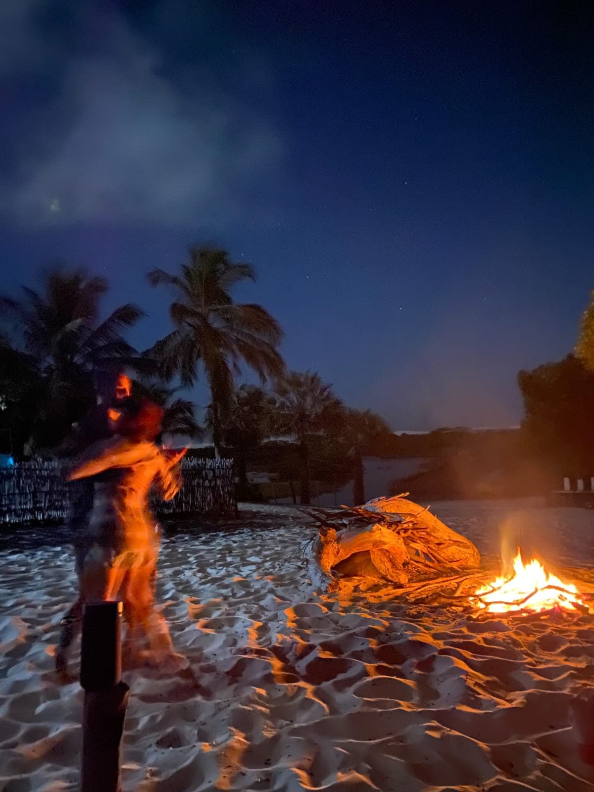 Warmer Sand, offenes Feuer, Sterne über den Palmen. Mehr braucht ein guter Abend nicht. Eine verwischte Silhouette steht neben einem knisternden Lagerfeuer auf weißem Sand bei Nacht während der Lençóis Maranhenses Wanderung, Palmen vor tiefblauem Sternenhimmel