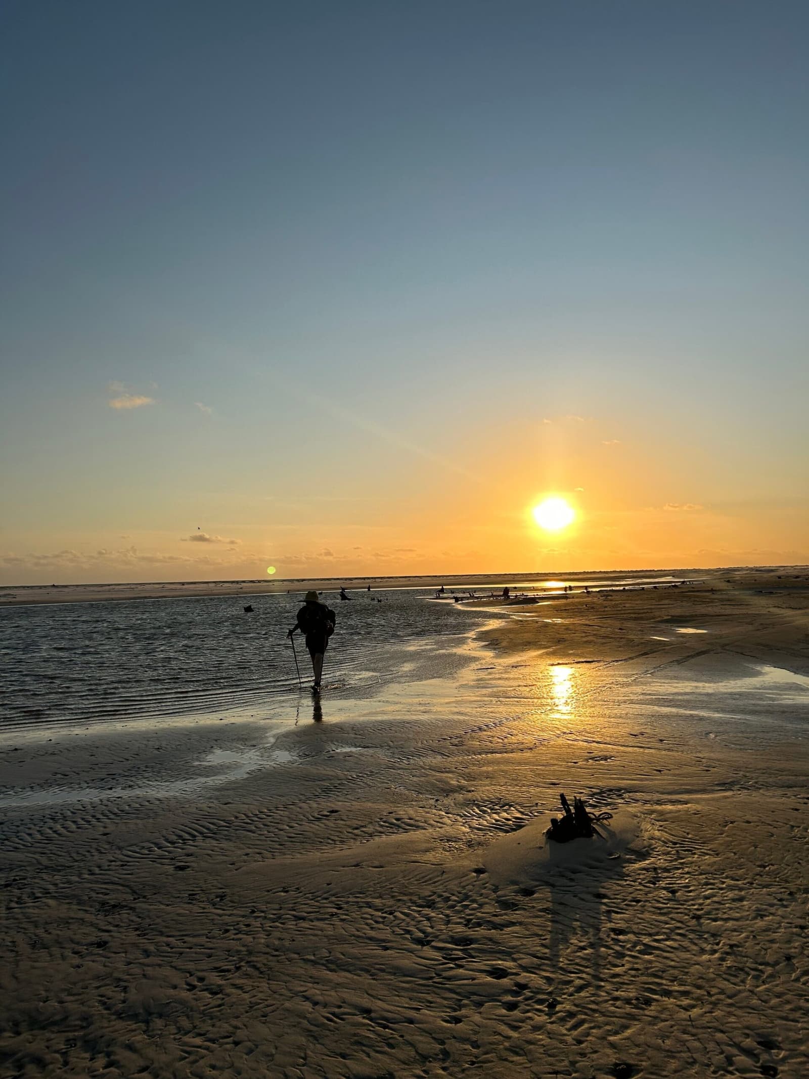 Allein auf den Salzflächen, die Sonne fast zum Greifen nah, so fühlt sich Weite an. Ein einsamer Wanderer mit Trekkingstöcken watet durch einen flachen Gezeitenkanal beim Lençóis-Maranhenses-Trek, als Silhouette vor dem goldenen Sonnenuntergang über den weitläufigen Sandwatt-Flächen