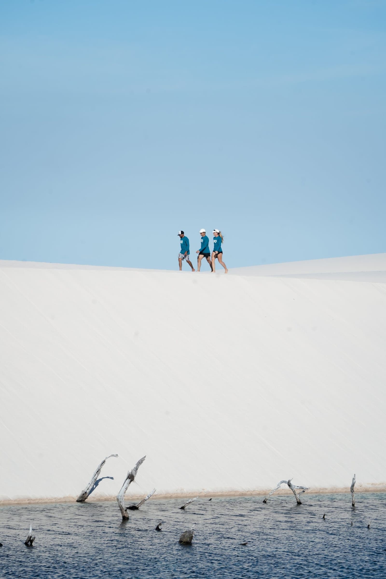 Oben auf der Düne wirkt selbst der Horizont weit weg. Drei Trekker in türkisfarbenen Shirts wandern entlang des Kamms einer weißen Sanddüne auf dem Lençóis-Maranhenses-Trek ab Atins, während im Vordergrund eine dunkle Lagune mit kahlen, im Wasser stehenden Ästen zu sehen ist