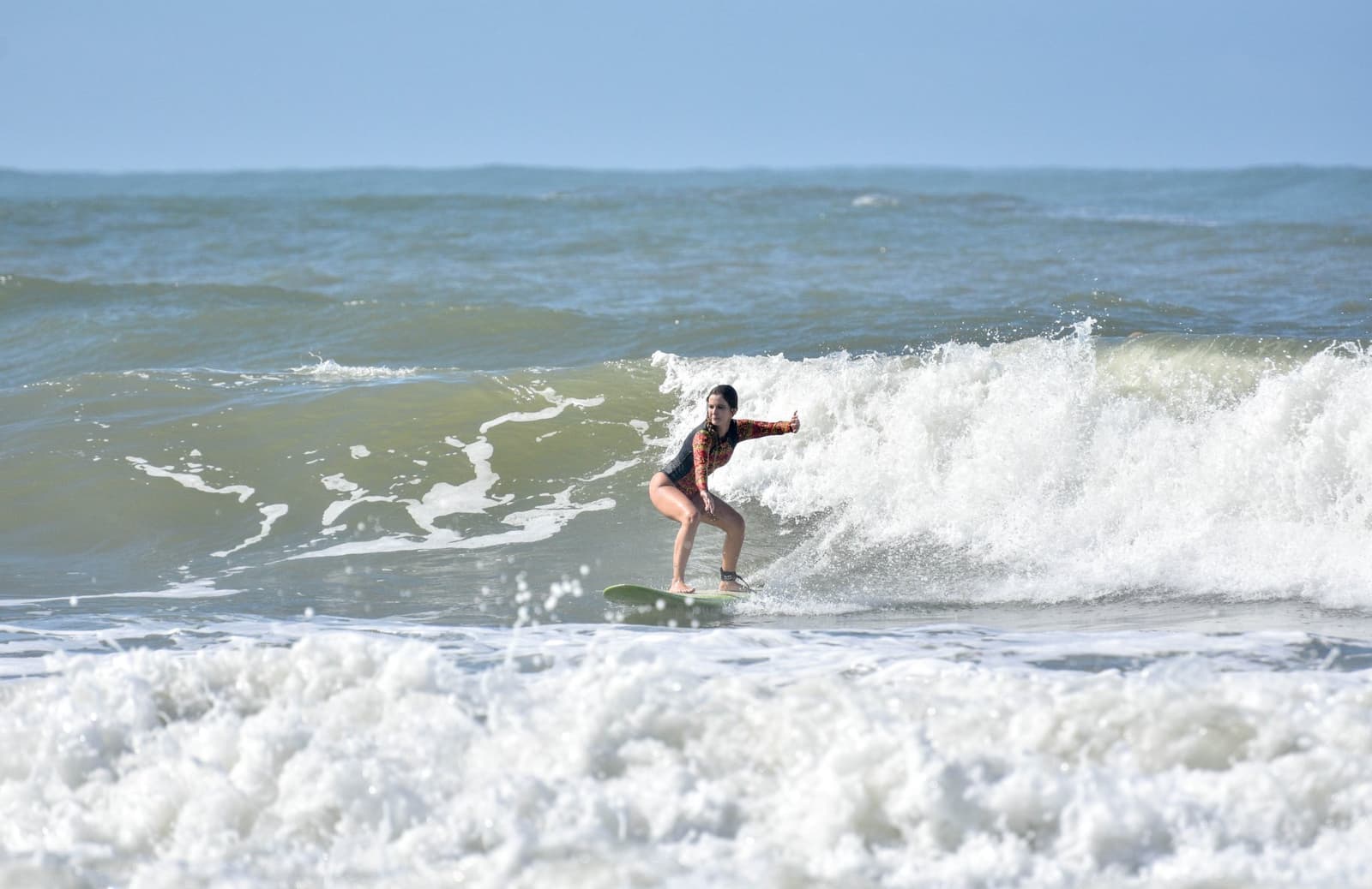 Braços abertos, olhar firme, a onda é dela. Photo: Picasa Uma mulher com rashguard de manga longa estampado cavalga uma onda quebrando durante aulas de surf em Itacaré, braços abertos para equilibrar sobre uma prancha verde