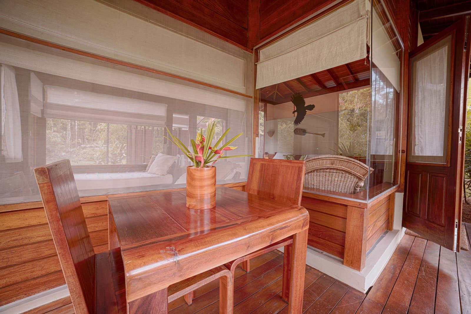 Le petit-déjeuner ici, après la sortie à l'aube, a un goût que les hôtels ordinaires ne connaissent pas. Photo: Samuel Melim Table et chaises en bois massif poli sur la véranda grillagée d'un bungalow de lodge d'observation des oiseaux en Amazonie, avec un arrangement de fleurs tropicales au premier plan et un lit de repos visible derrière la moustiquaire