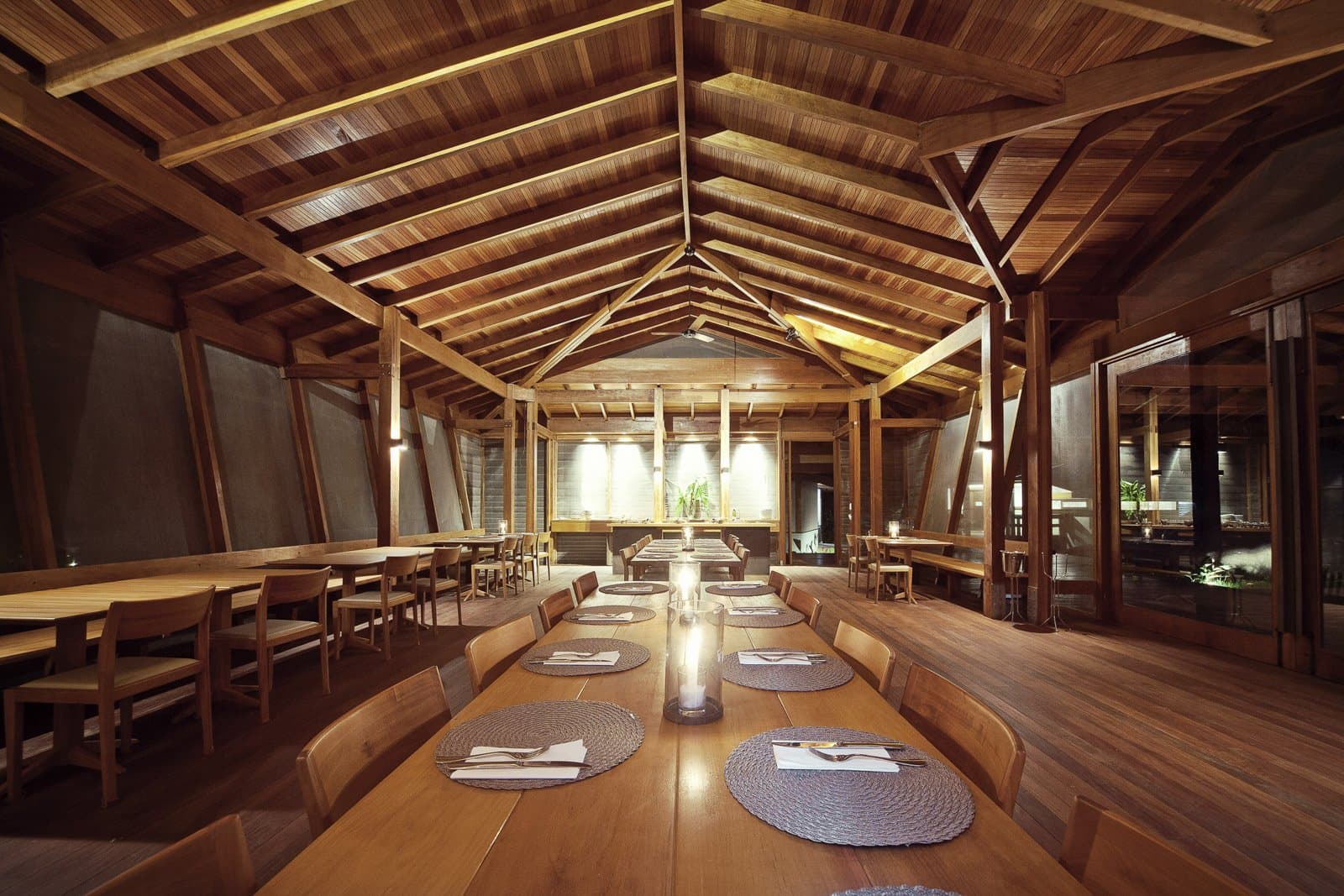 Grande table, bonne compagnie, et la forêt à deux pas derrière la vitre. Photo: Samuel Melim (c) Salle à manger éclairée aux bougies dans un lodge de birdwatching en forêt amazonienne, avec une longue table communautaire en bois, des sets de table tressés et une charpente en bois apparente