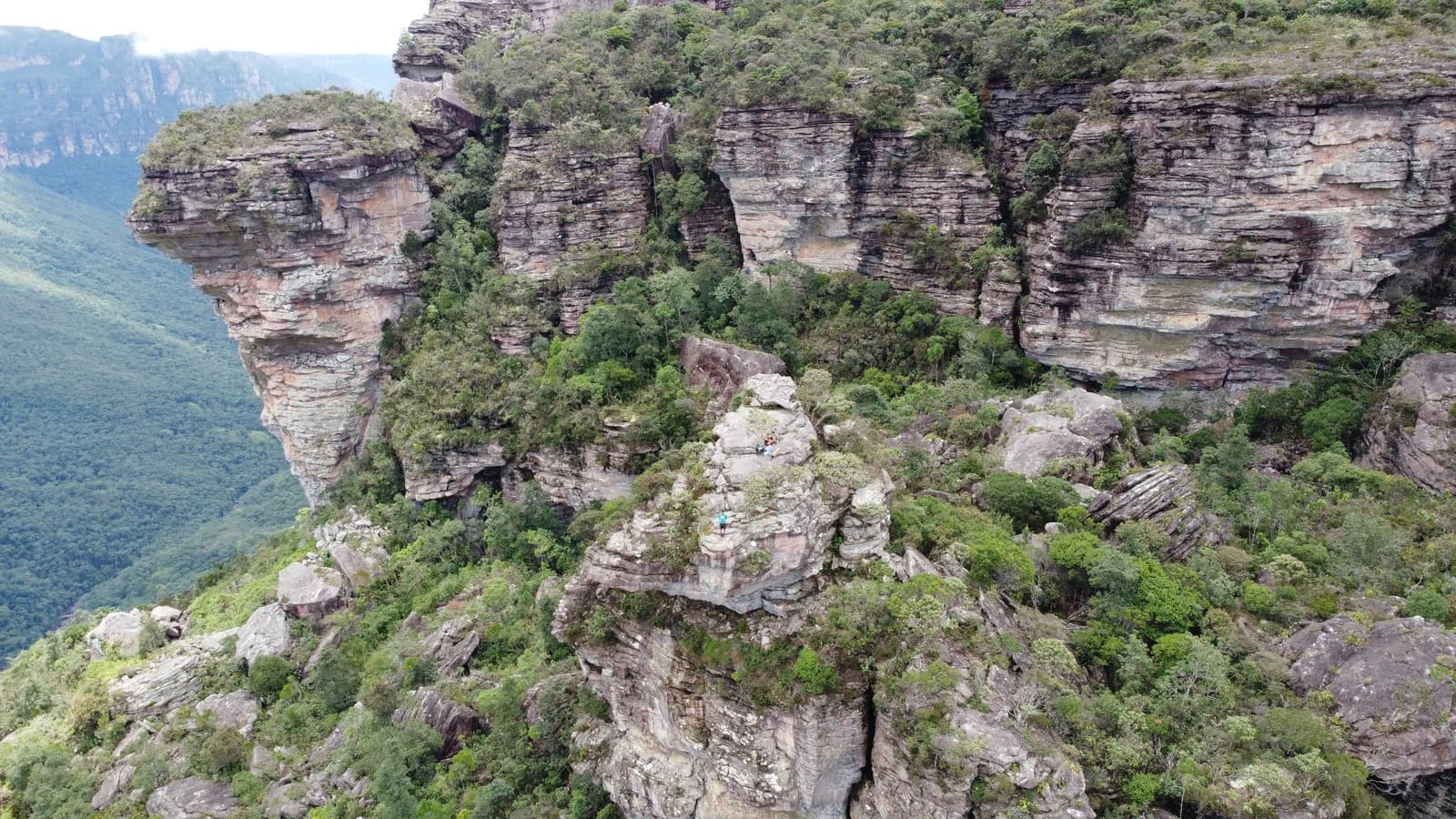 Zwei Punkte auf einem Felsvorsprung, darunter ein ganzes Canyon-Tal. Luftaufnahme von geschichteten Sandsteinklippen und grüner Vegetation im Chapada-Diamantina-Trek, mit zwei winzigen Wanderern auf einem freistehenden Felsvorsprung über einem tiefen Canyon
