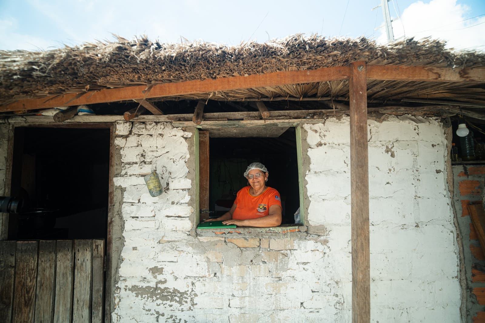 Kein Schild nötig, dieser Blick sagt alles. Eine lächelnde Frau mit Haarnetz und orangefarbenem T-Shirt lehnt aus einem kleinen Fenster eines weiß getünchten Lehmhauses mit Palmstrohdach – Teil einer Gastfamilien-Unterkunft auf dem Lençóis-Maranhenses-Trek ab Atins.