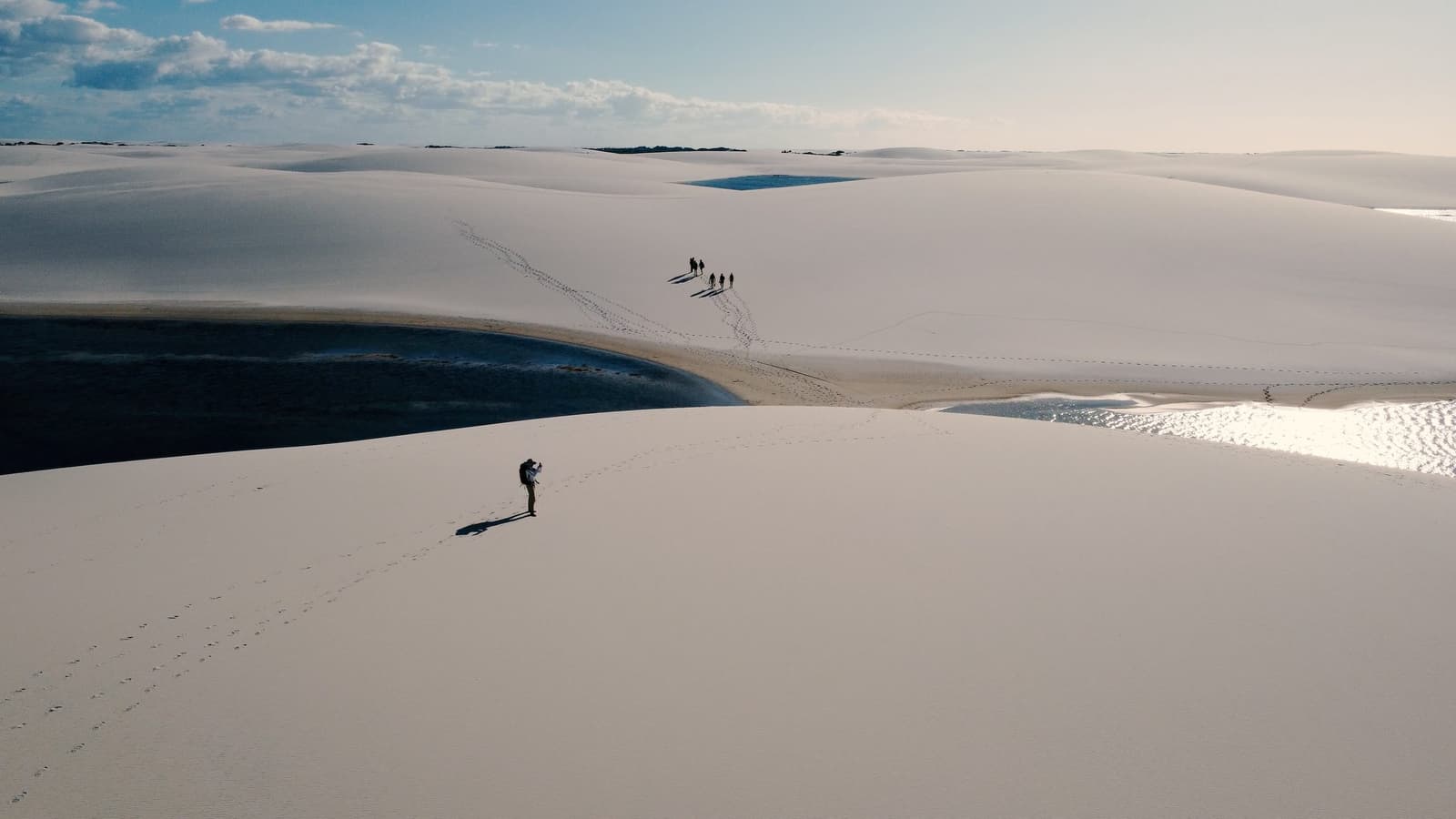 On se sent petit, et c'est exactement ça qui fait du bien. Vue aérienne d'un photographe solitaire debout sur les immenses dunes de sable blanc lors d'un trekking dans les Lençóis Maranhenses, avec des lagunes aux eaux sombres et un petit groupe de randonneurs visible au loin