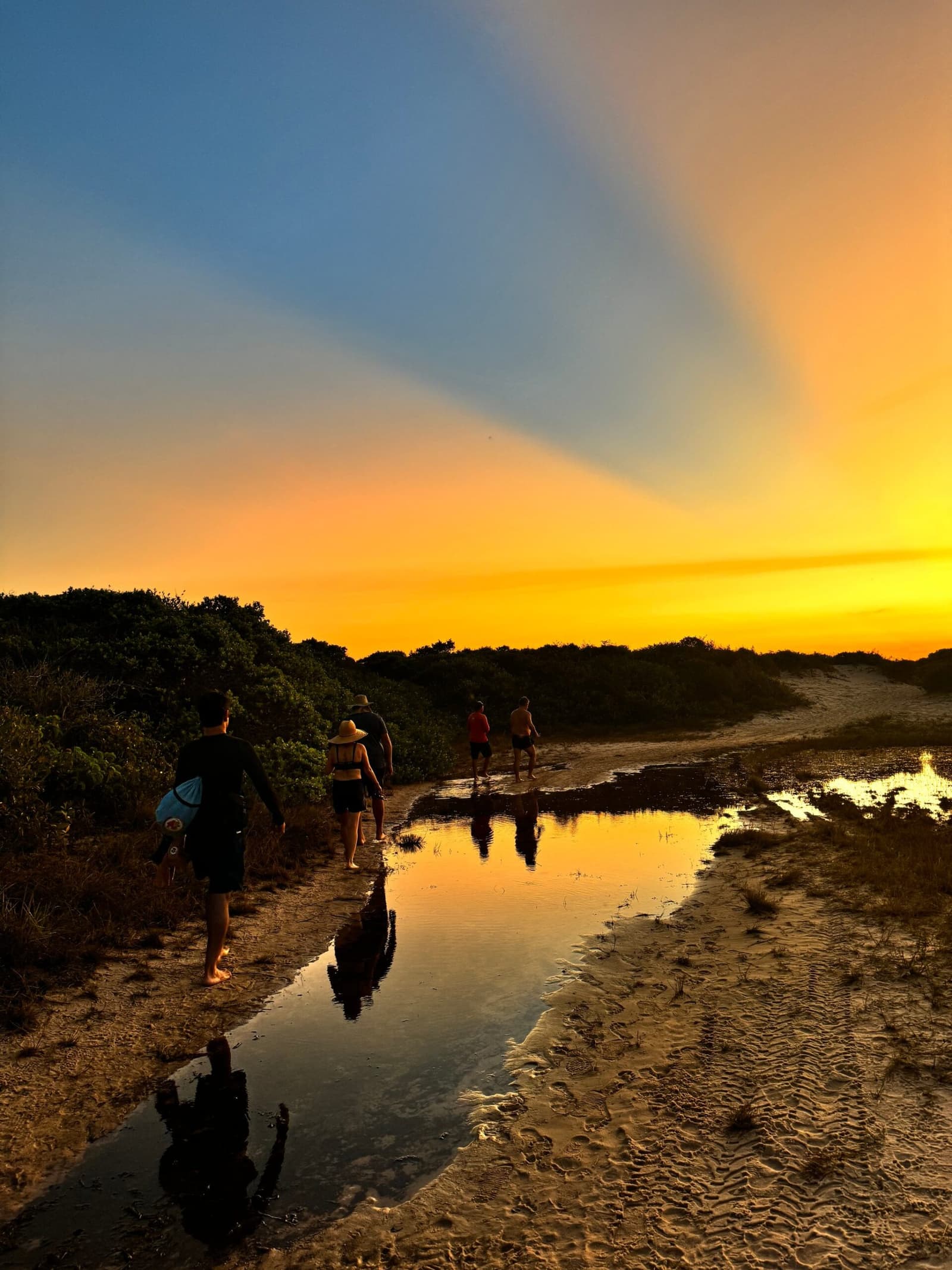 Ceux qui ont continué à marcher ont eu le meilleur spectacle du soir. Cinq randonneurs marchent pieds nus sur un sentier sablonneux lors d'un trekking dans les Lençóis Maranhenses, leurs silhouettes et le ciel doré du coucher de soleil se reflétant dans un mince cours d'eau