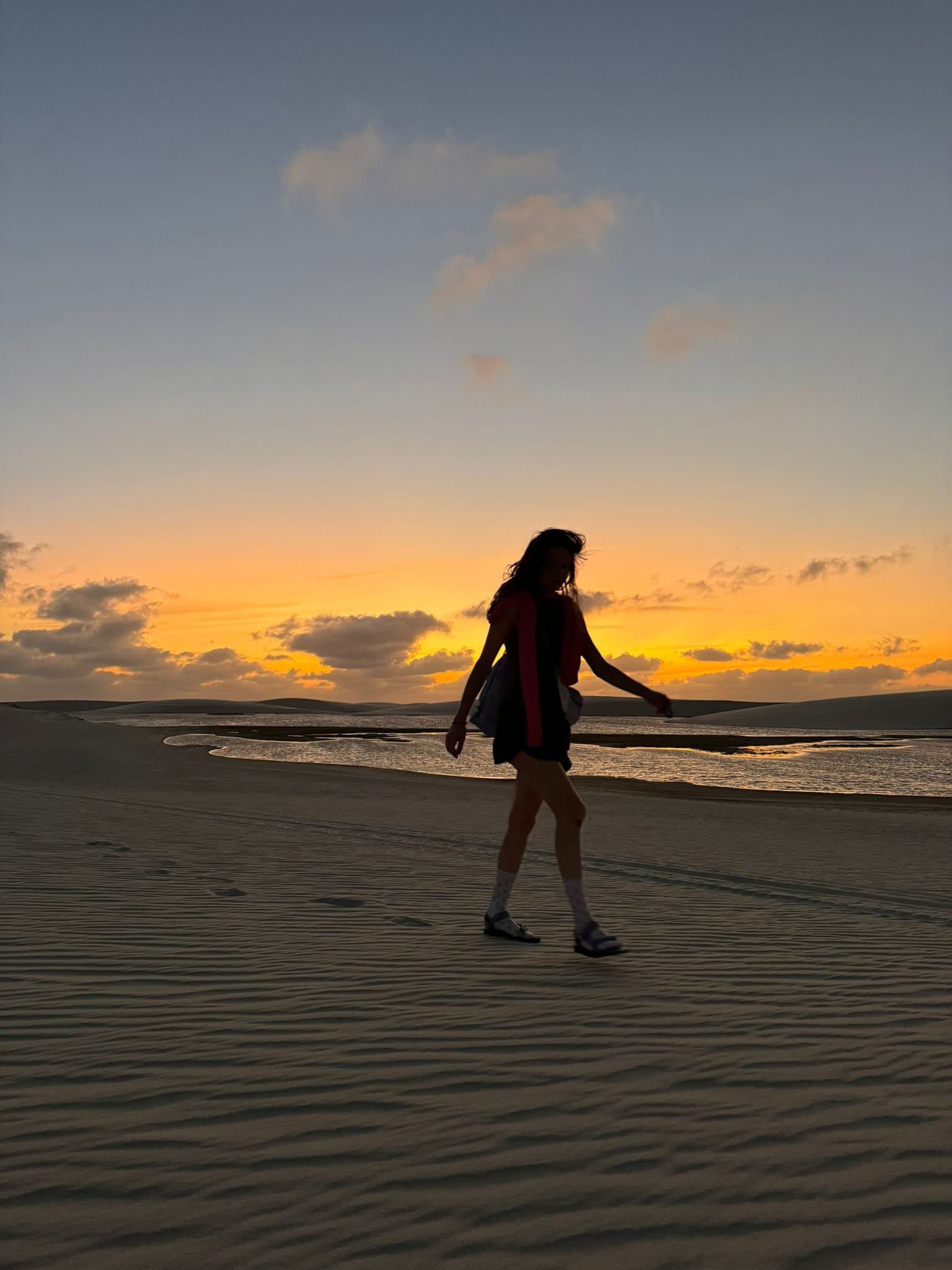 Sand, Wind und ein Horizont, der einem gehört. Silhouette einer Frau, die beim Lençóis-Maranhenses-Trekking über gewellte Sanddünen läuft, während ein orangefarbener Sonnenuntergang und eine flache Lagune den Himmel spiegeln