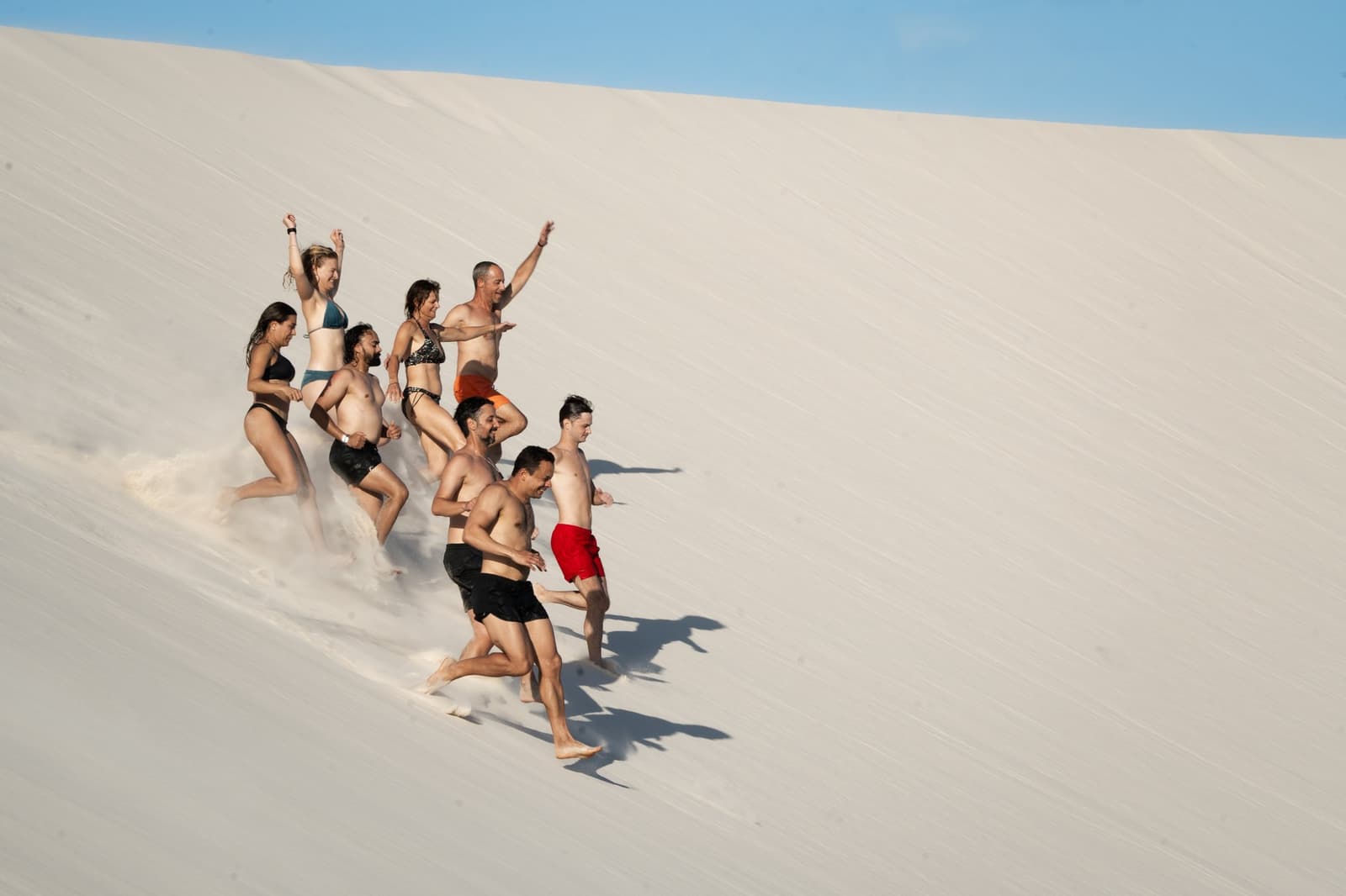 Eine Düne hinunterrennen: schneller als gedacht, lustiger als erwartet. Eine Gruppe von acht Reisenden in Badekleidung läuft lachend eine steile weiße Sanddüne beim Lençóis-Maranhenses-Trek ab Atins hinunter, mit erhobenen Armen und aufgewirbeltem Sand unter blauem Himmel.