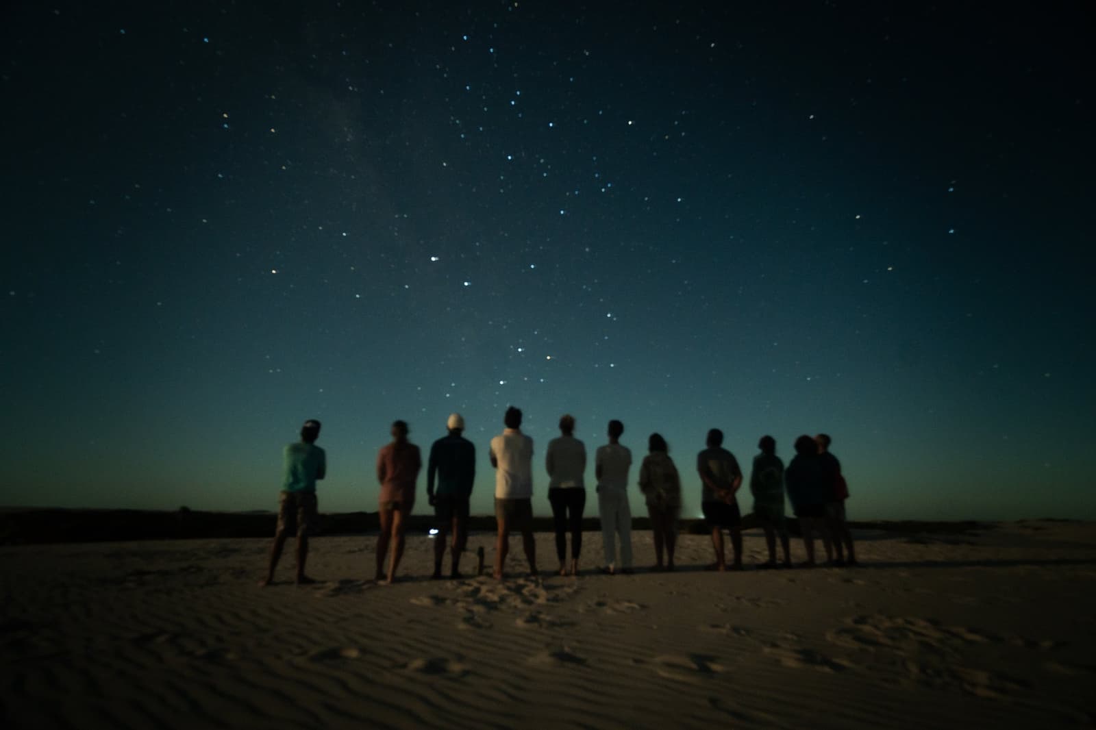 No light pollution, no distractions, just ten people and the whole sky. A group of ten people standing in a row on open sand dunes, silhouetted against a vast star-filled night sky during a lencois maranhenses trek