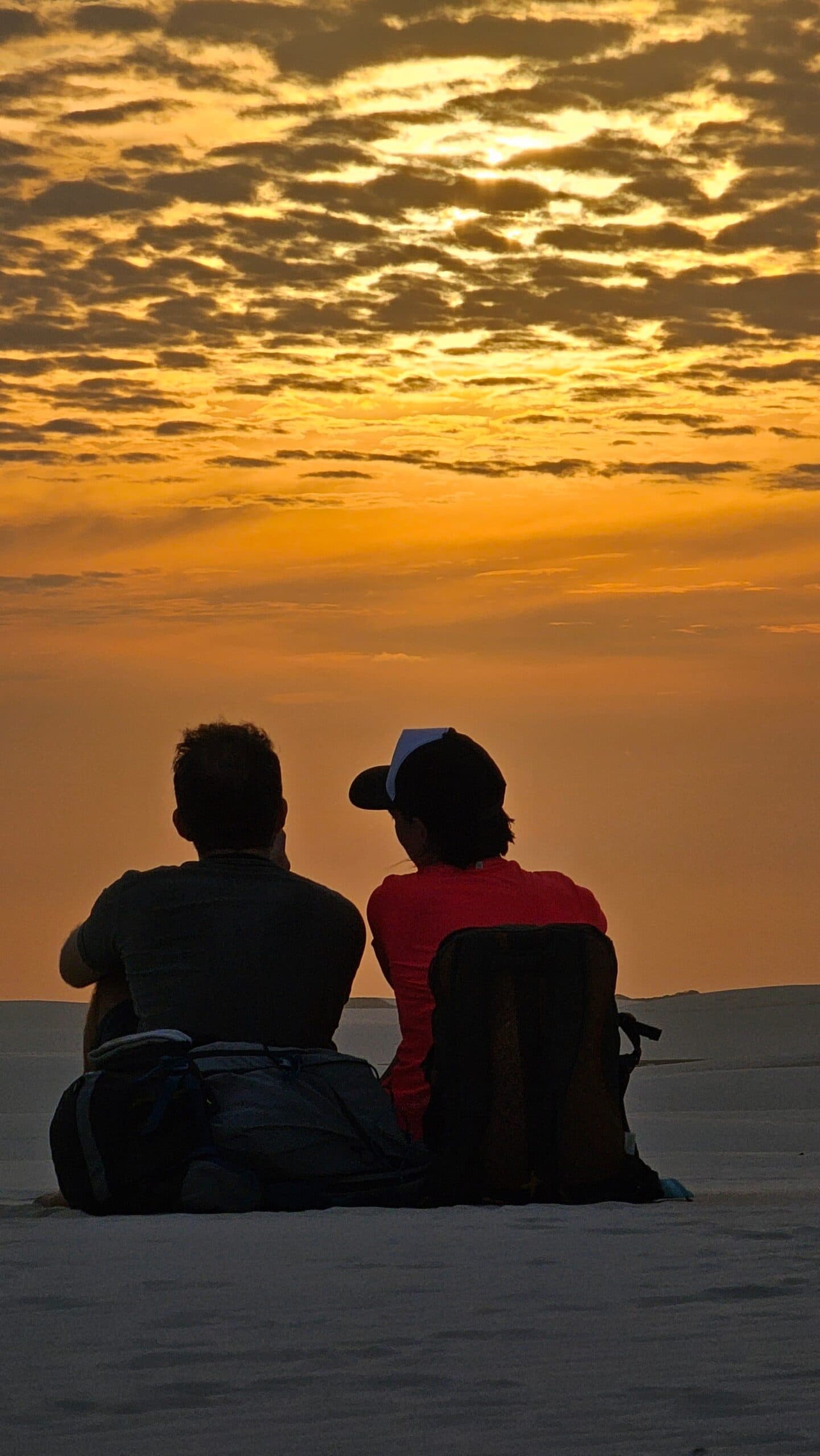 Plus de réseau, plus d'horaires, juste ce ciel incandescent. Deux randonneurs assis côte à côte sur les dunes de sable blanc du trek des Lençóis Maranhenses, en silhouette sur un ciel doré et orangé parsemé de nuages