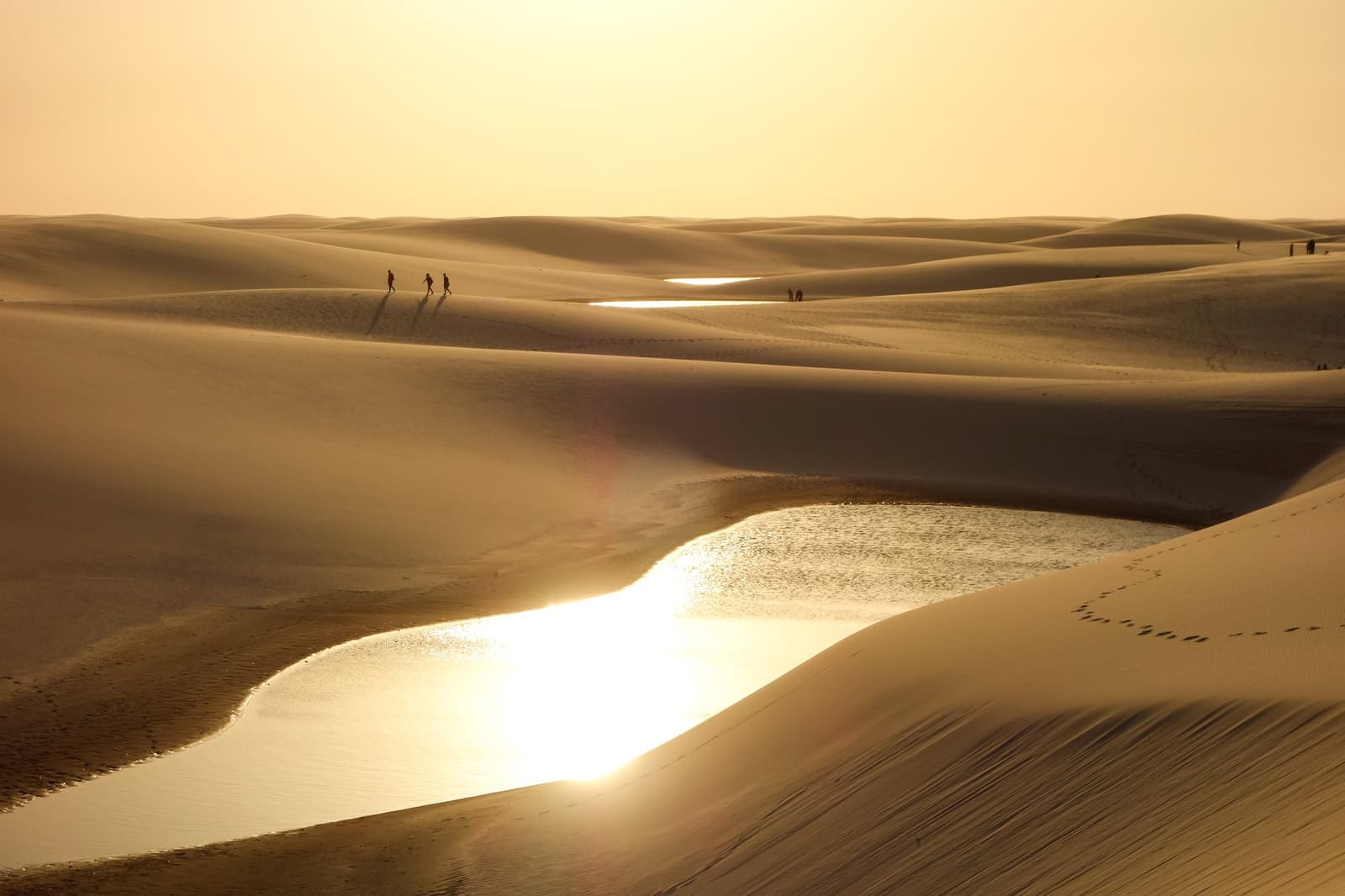 Eine Wüste, die Wasser sammelt. Kleiner Spoiler: Es lohnt sich. Drei Silhouetten wandern beim Lençóis Maranhenses Hike über einen goldenen Dünenkamm, während flache Regenwasserlagunen im tiefen Abendlicht über das weite Dünenfeld glitzern