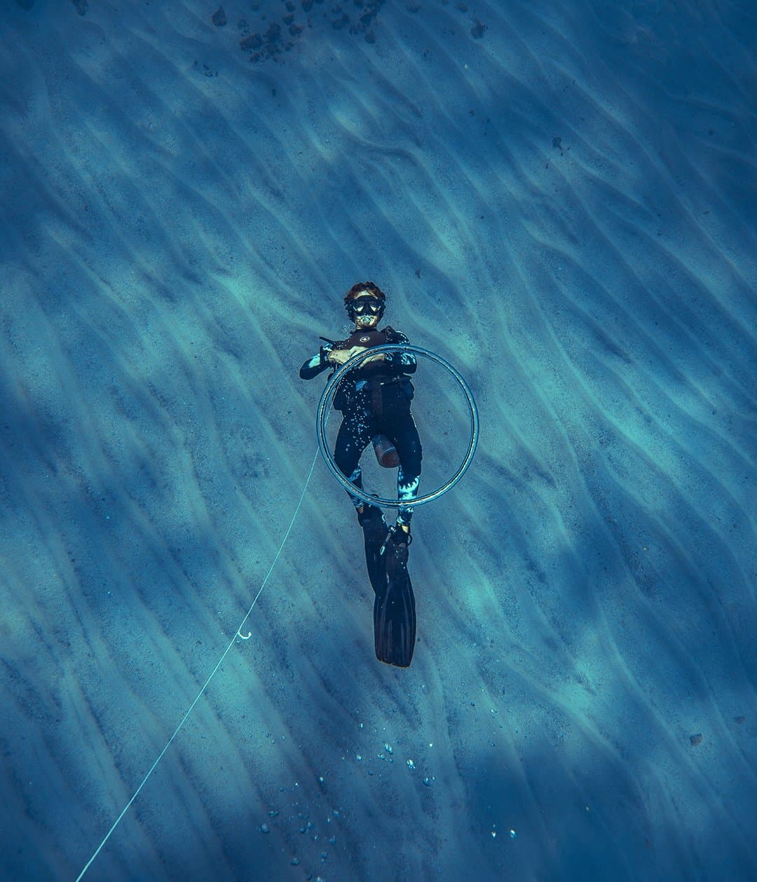 Just a person, a coil of line, and a whole lot of blue below. Aerial view of a scuba diver in a black wetsuit holding a large ring, suspended above a rippled sandy seabed during the 2-Day Diving Package in Fernando de Noronha