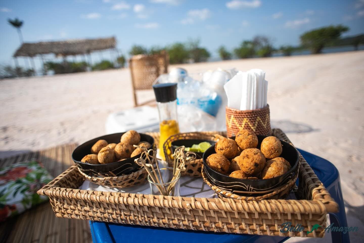 Sandbank-Pause mit Knabbereien, die beweisen: Der Amazonas kennt auch Genuss. Ein geflochtenes Rattantablett mit zwei schwarzen Schalen voller goldener frittierter Snackbällchen, serviert am Sandstrand während einer Amazonas-Flusskreuzfahrt ab Alter do Chão, mit Bambusspießen, Limettenspalten und Gewürzen