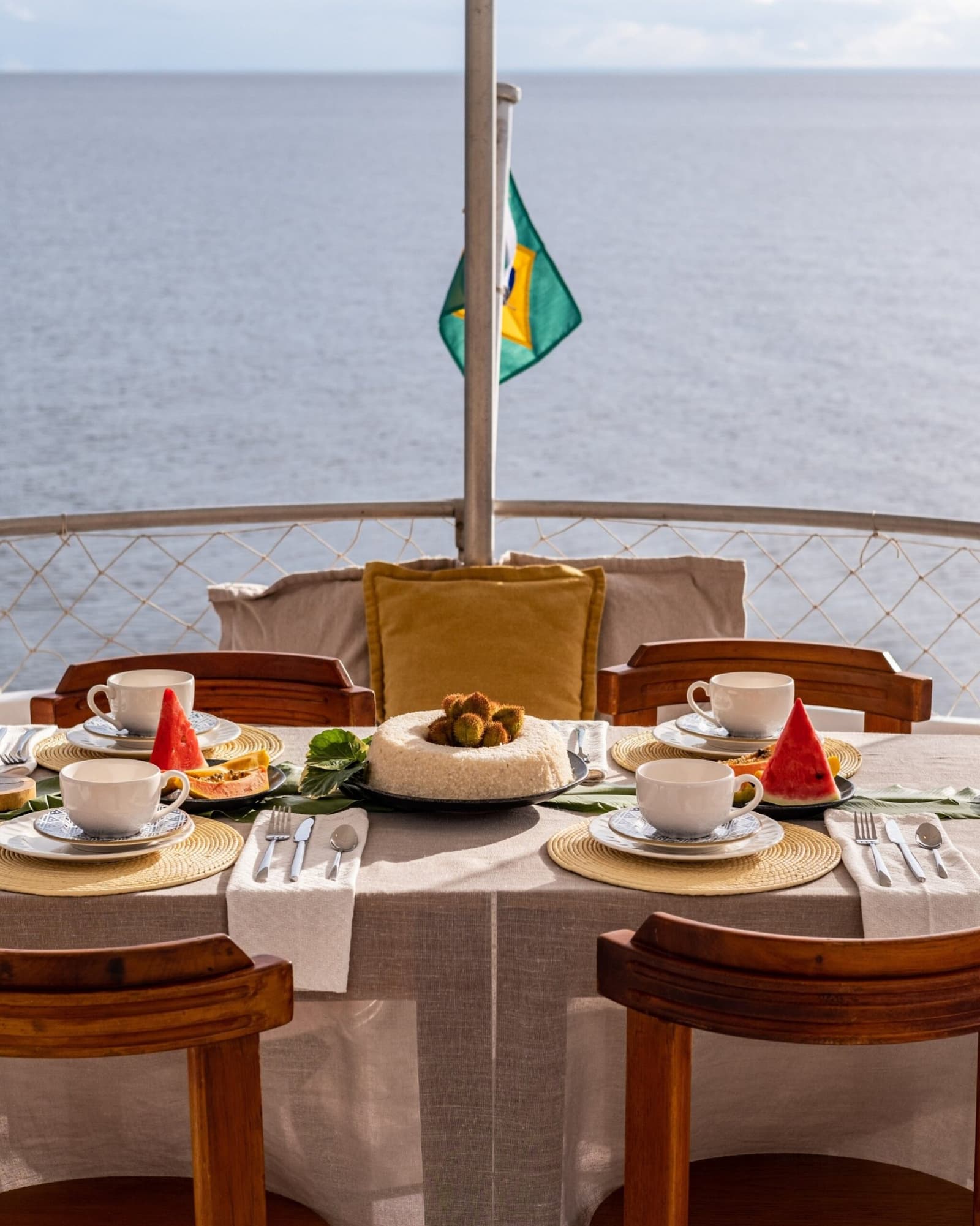 Frühstück mit Flussblick, der beste Grund, früh aufzustehen. Gedeckter Frühstückstisch auf dem offenen Deck einer Amazonas-Kreuzfahrt ab Alter do Chão, mit frischer Wassermelone, Papaya, weißen Kaffeetassen und einem Mittelstück-Kuchen, brasilianische Flagge über ruhigem Wasser im Hintergrund