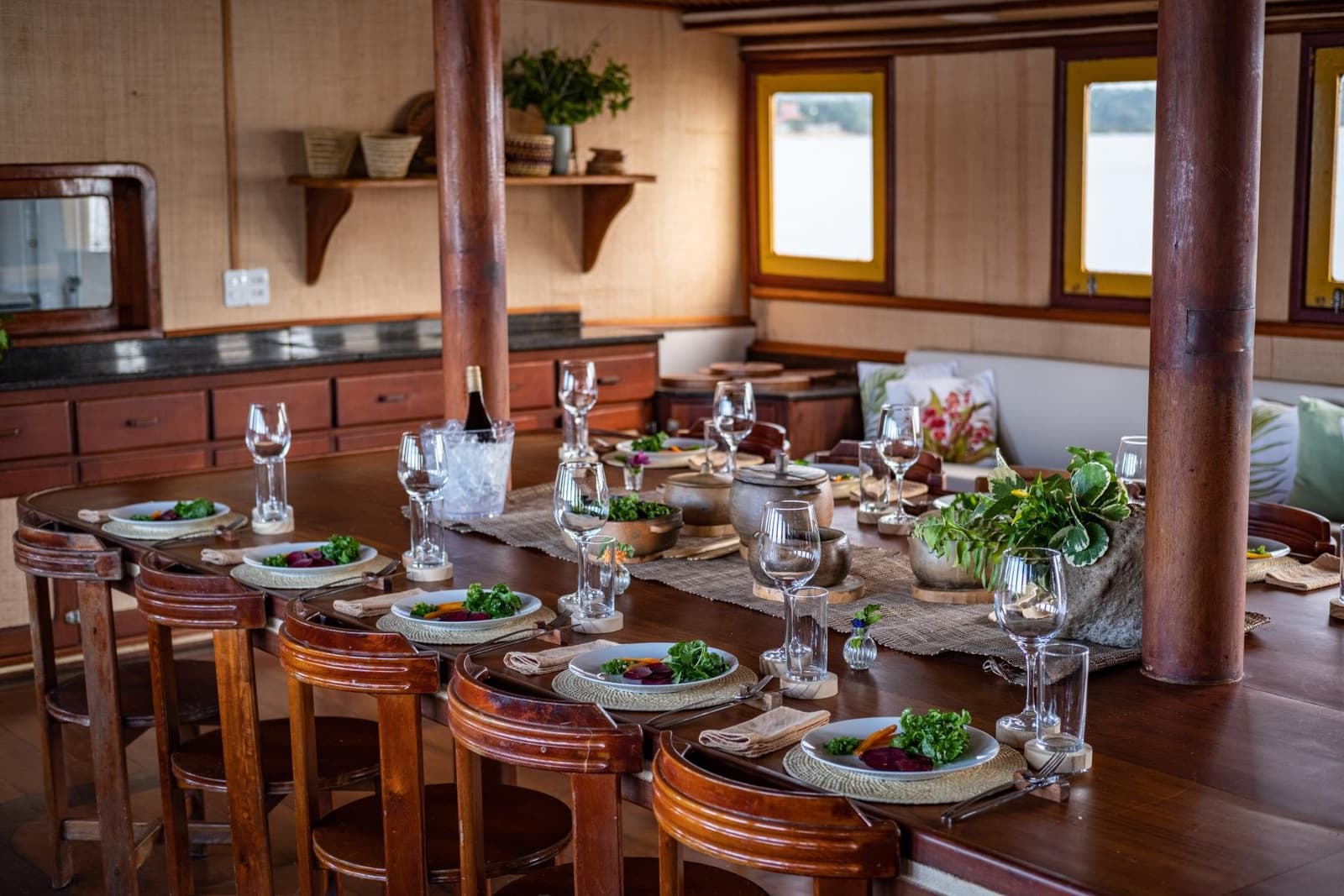 Frische Küche, Flussluft und ein Tisch, der auf Sie wartet. Elegant gedeckter ovaler Esstisch auf einem Amazonas-Kreuzfahrtschiff von Alter do Chão, mit frischen Salatplatten, Weingläsern, einer Flasche im Eiskühler, rustikalen Holzschemeln und Flussblick durch gelbgerahmte Bullaugen