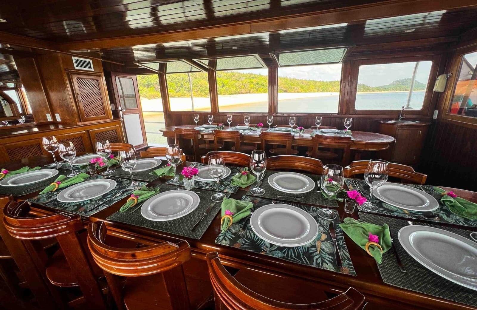 Abendessen mit Flussblick, so sieht ein guter Abend auf dem Amazonas aus. Elegant gedeckte Esstische mit Weingläsern, grünen Servietten und rosa Blütendekor im polierten Holzsalon einer Amazon-Kreuzfahrt von Alter do Chão, mit Blick auf einen Flussstrand und bewaldete Hügel durch Panoramafenster
